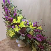 Tall asymmetrical arrangement of yellow lilies and purple flowers in a white pedestal vase