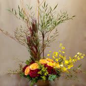 Tall autumn-toned arrangement with orange roses, red blooms, yellow flowers and branches in a brown vase