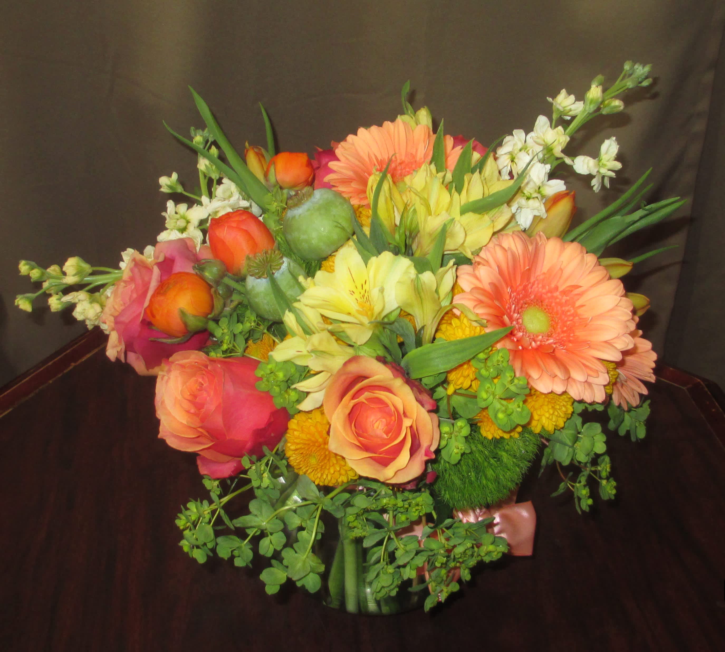 Low glass vase of orange gerberas, roses, and mixed yellow flowers in a compact arrangement