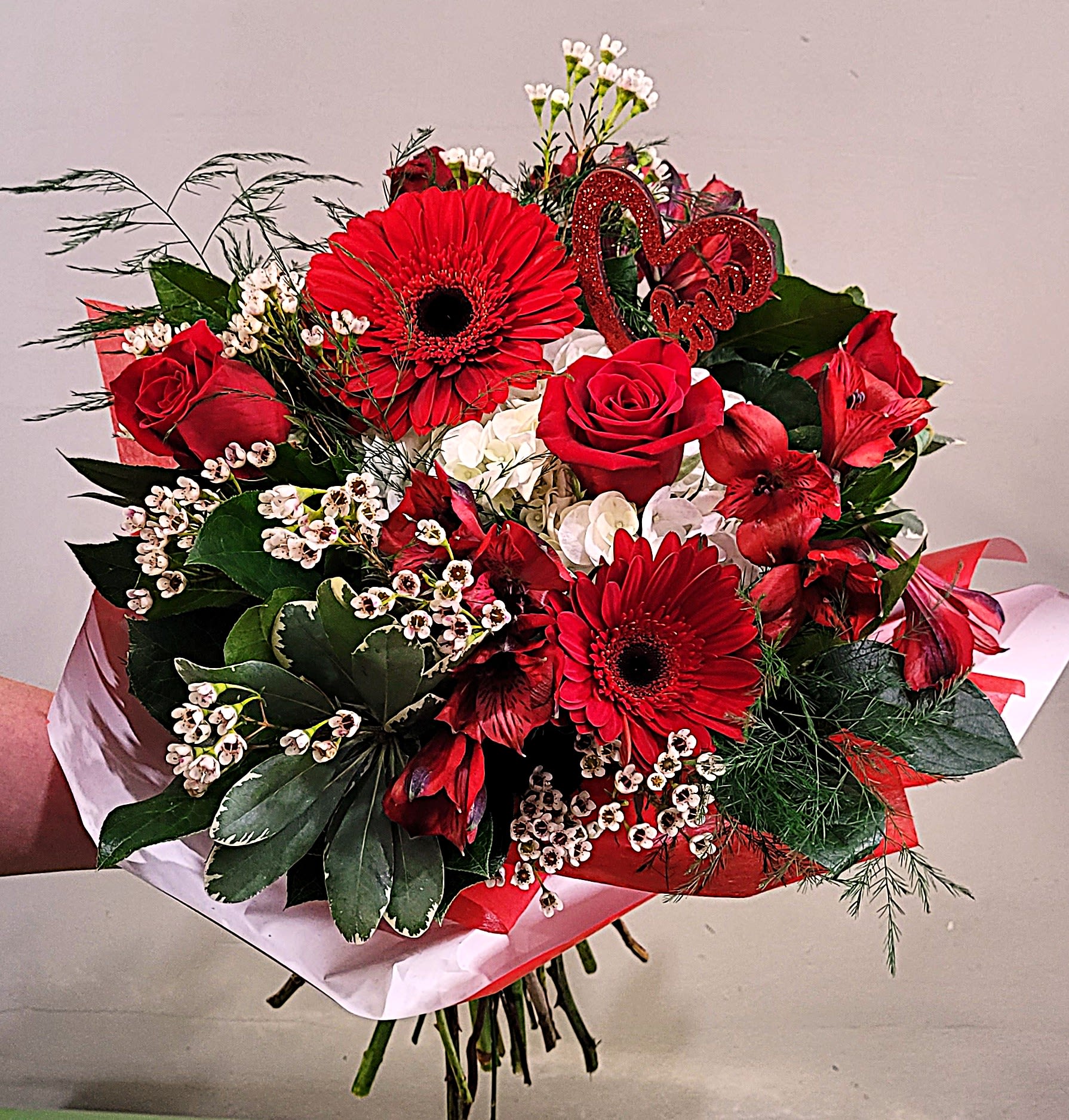 Hand-tied bouquet of red roses and gerbera daisies with white accents and a red heart pick