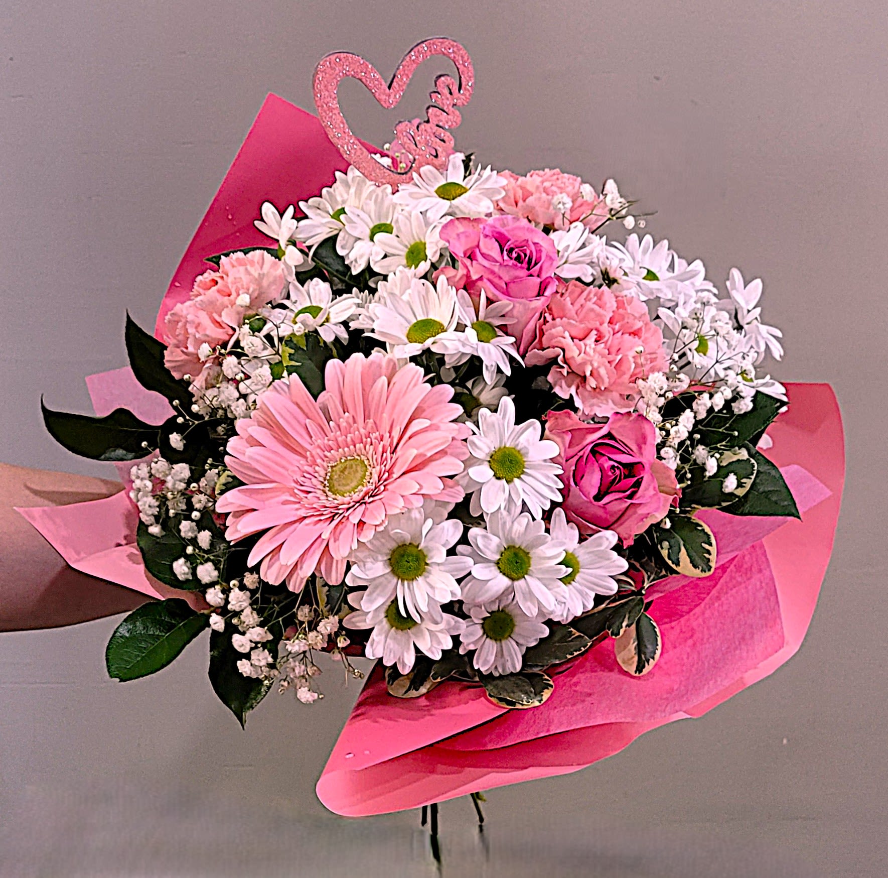 Hand-tied bouquet of pink gerbera, roses, and carnations with white daisies wrapped in pink paper and a heart pick.