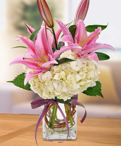 Pink lilies and white hydrangeas in a clear glass vase with a purple ribbon