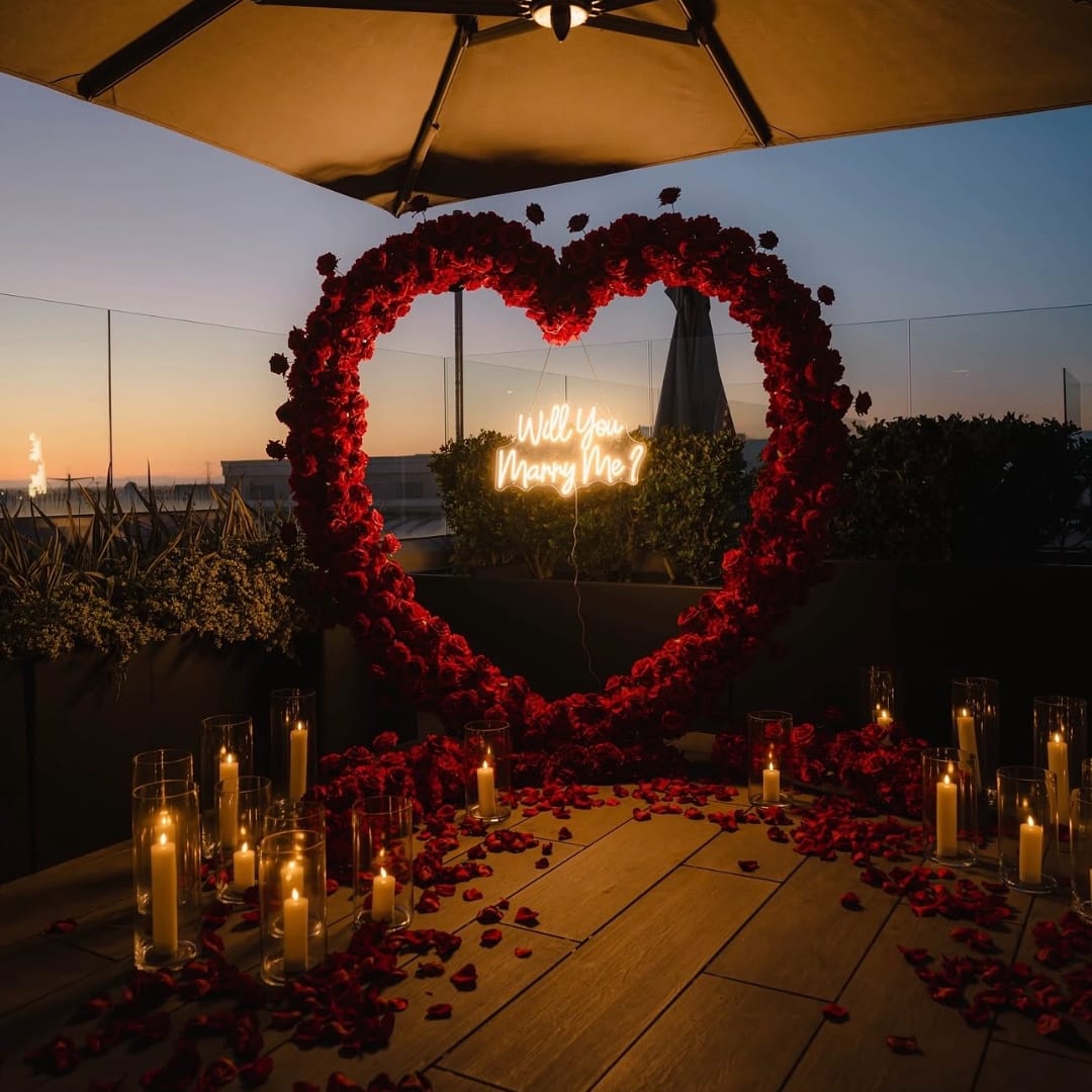 Heart-shaped red floral arch with candles and a neon "Will You Marry Me?" sign