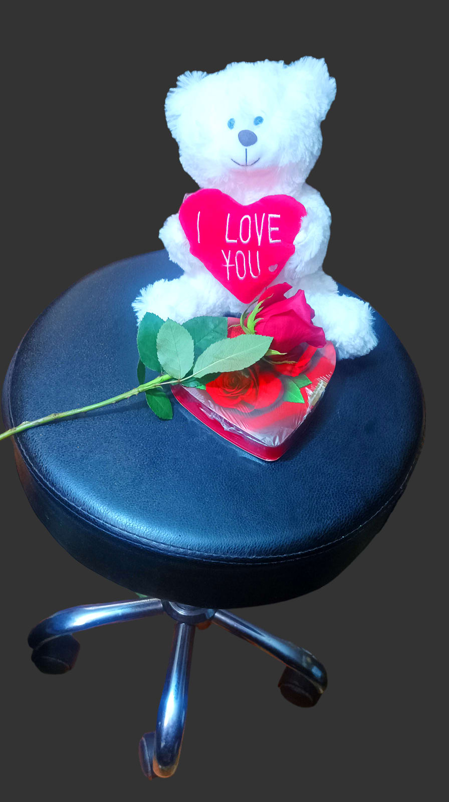White teddy bear holding a pink heart with a red rose on a stool