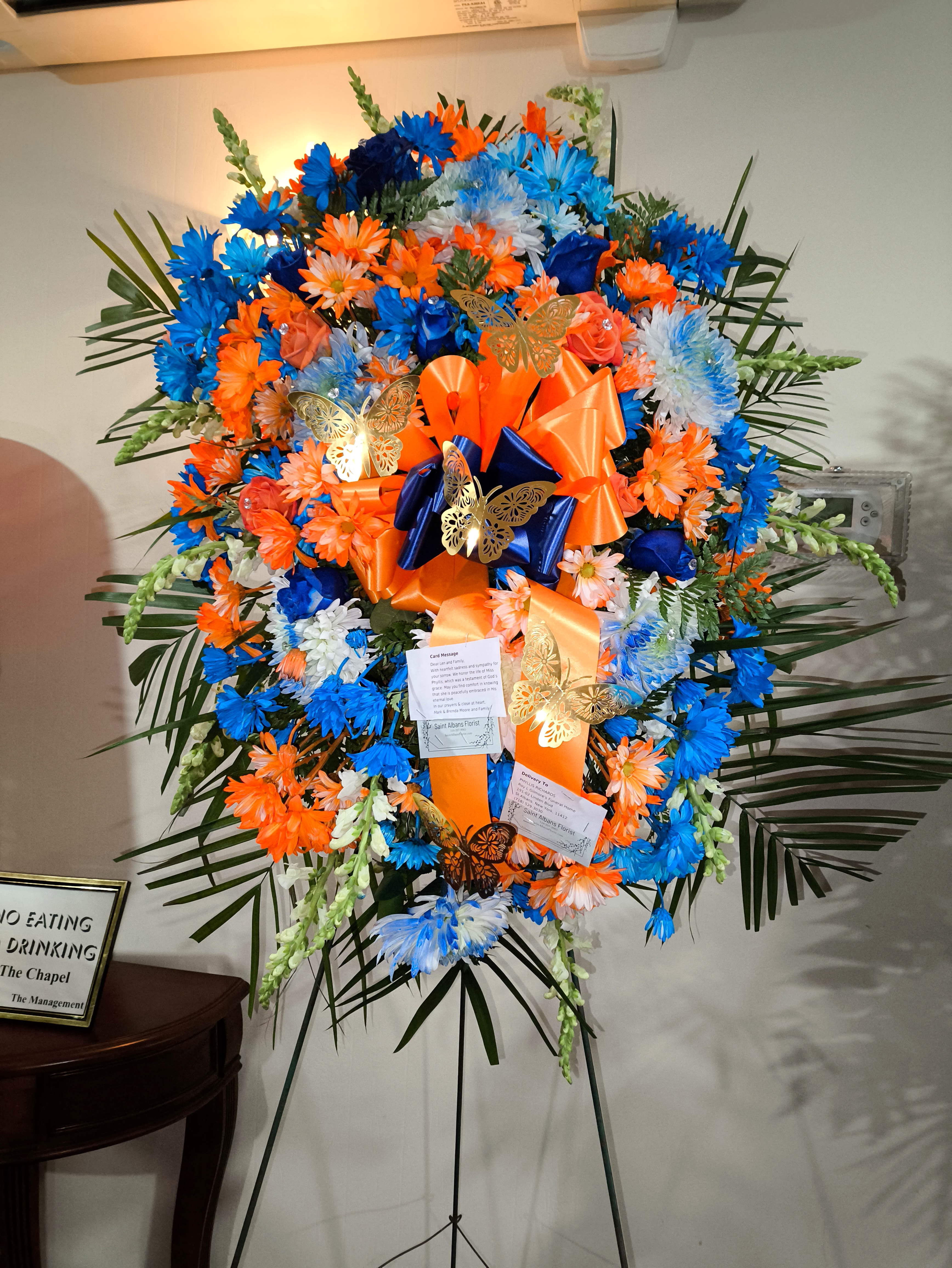 Large standing floral spray in blue, orange, and white with ribbons and butterflies