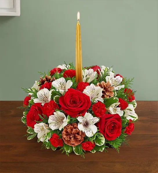 Christmas Carol Centerpiece - Our festive centerpiece brings a classic touch to all your holiday celebrations. We’ve gathered a mix of red and white blooms with lush greens and rustic pinecones. Gold taper candles add a warm and cozy Christmas glow. Type of white blooms may vary from carnation to alstromeria to stock. 