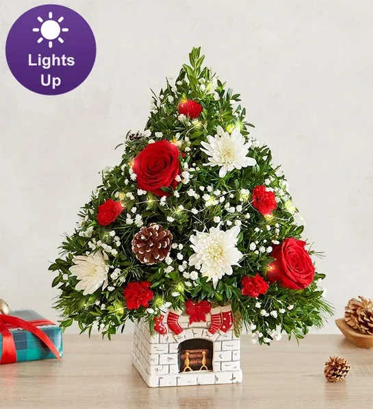 Holiday Tree Merry Mantel - The joy of gift-giving comes alive in the latest edition of our iconic Holiday Flower Tree, trimmed in festive blooms. It’s designed in our exclusive “The Stockings were Hung Mantel”, which captures the anticipation of Santa’s arrival. This white ceramic container is decorated with four stockings, an evergreen garland and a festive red bow. Complete with white-tipped pinecones and a warm white fairy lights, it’s a magical collectible for the holiday season.