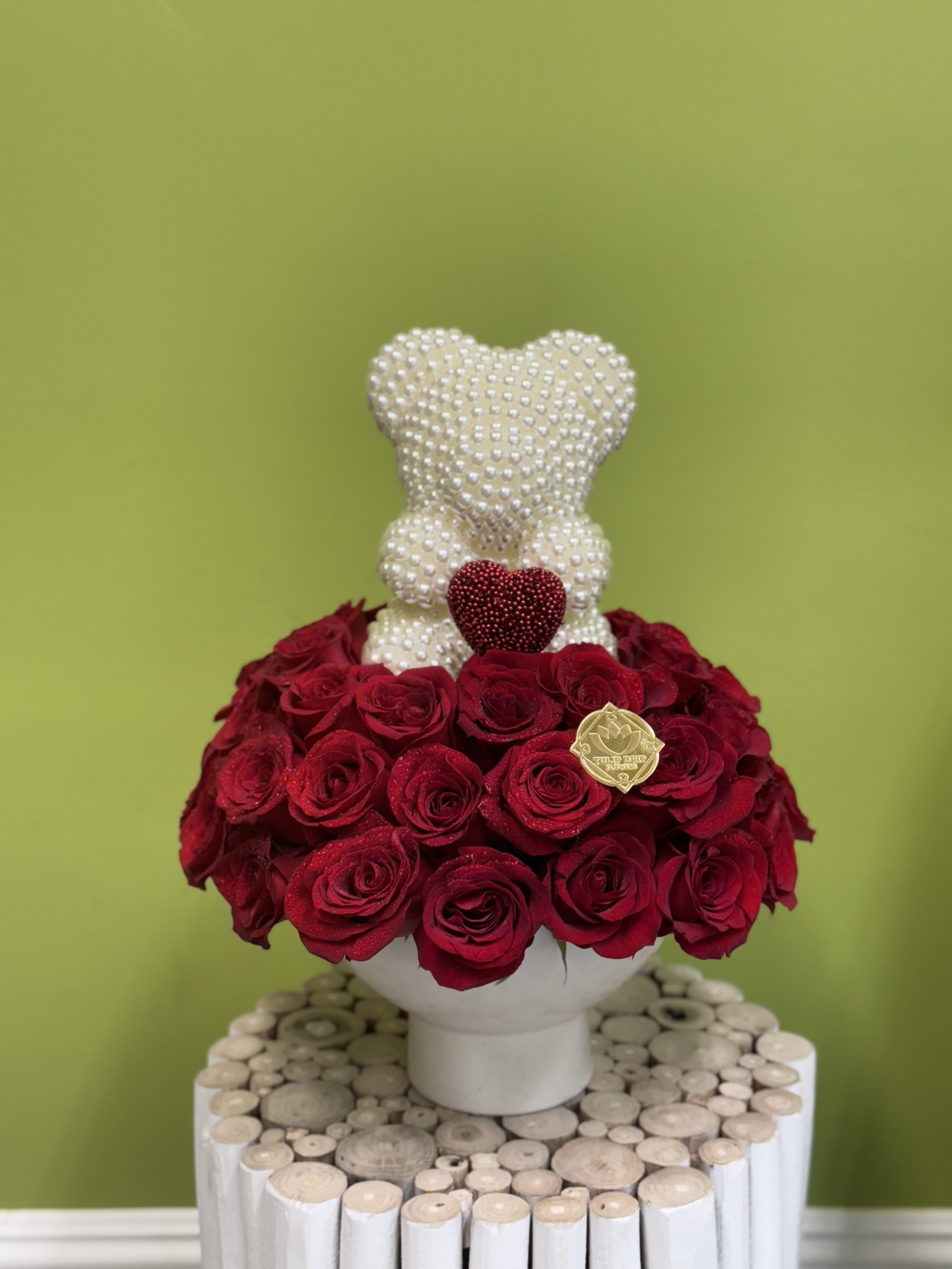 Red roses arranged around a pearl-covered teddy bear in a white vase