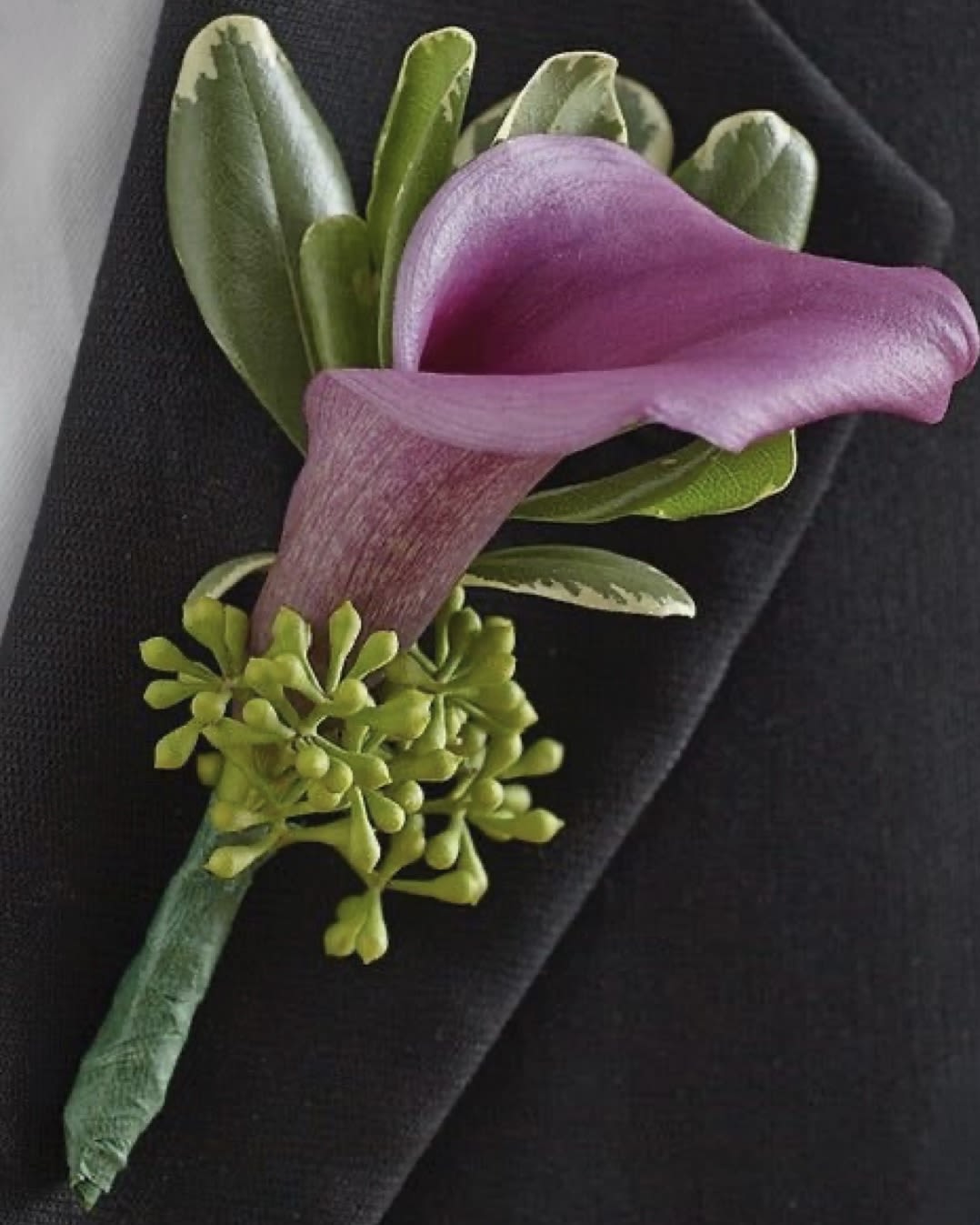 Purple calla lily boutonniere with green accents on a dark suit lapel