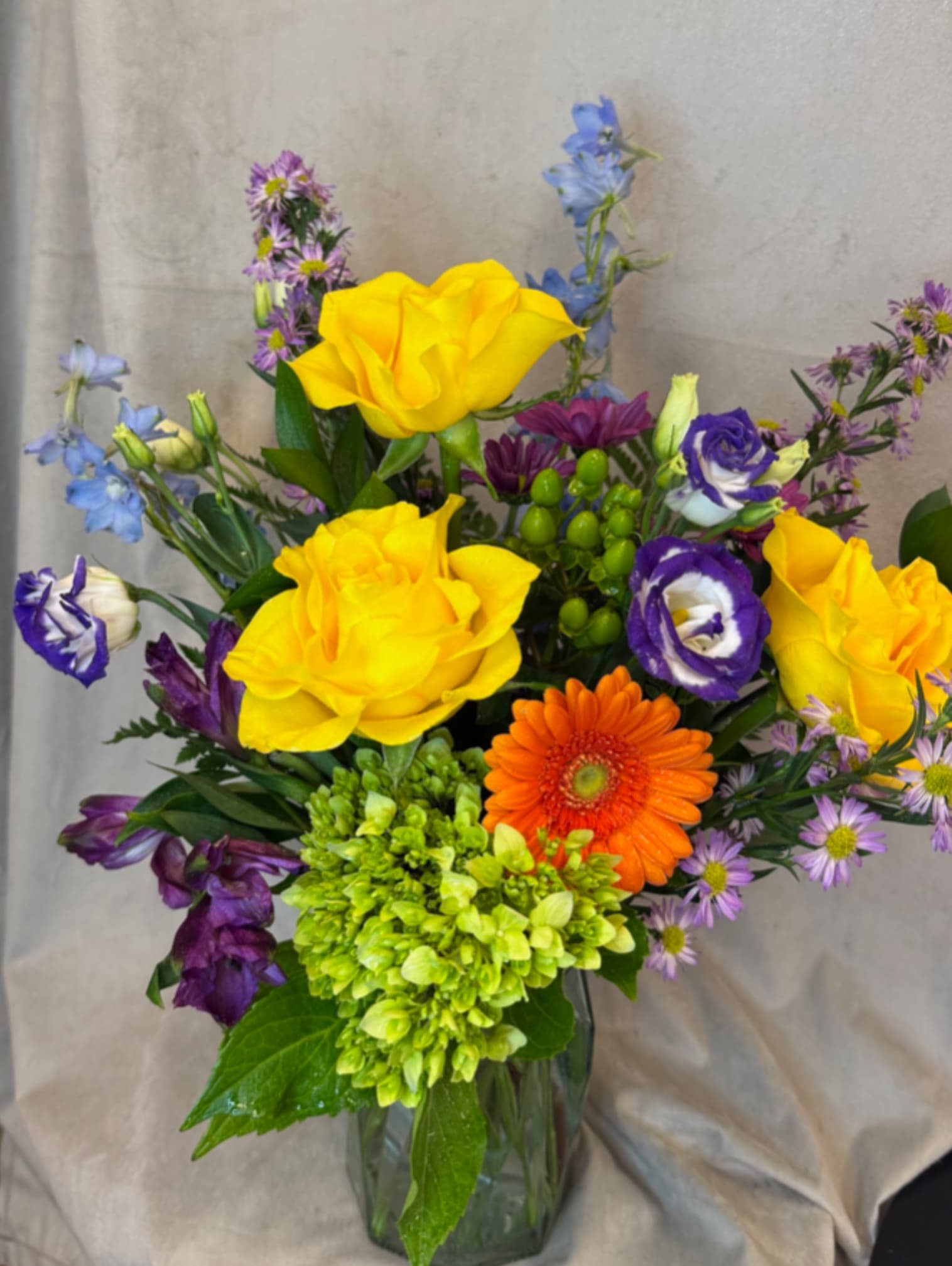 Mixed bouquet with yellow roses, orange gerbera daisy, and purple blooms in a glass vase