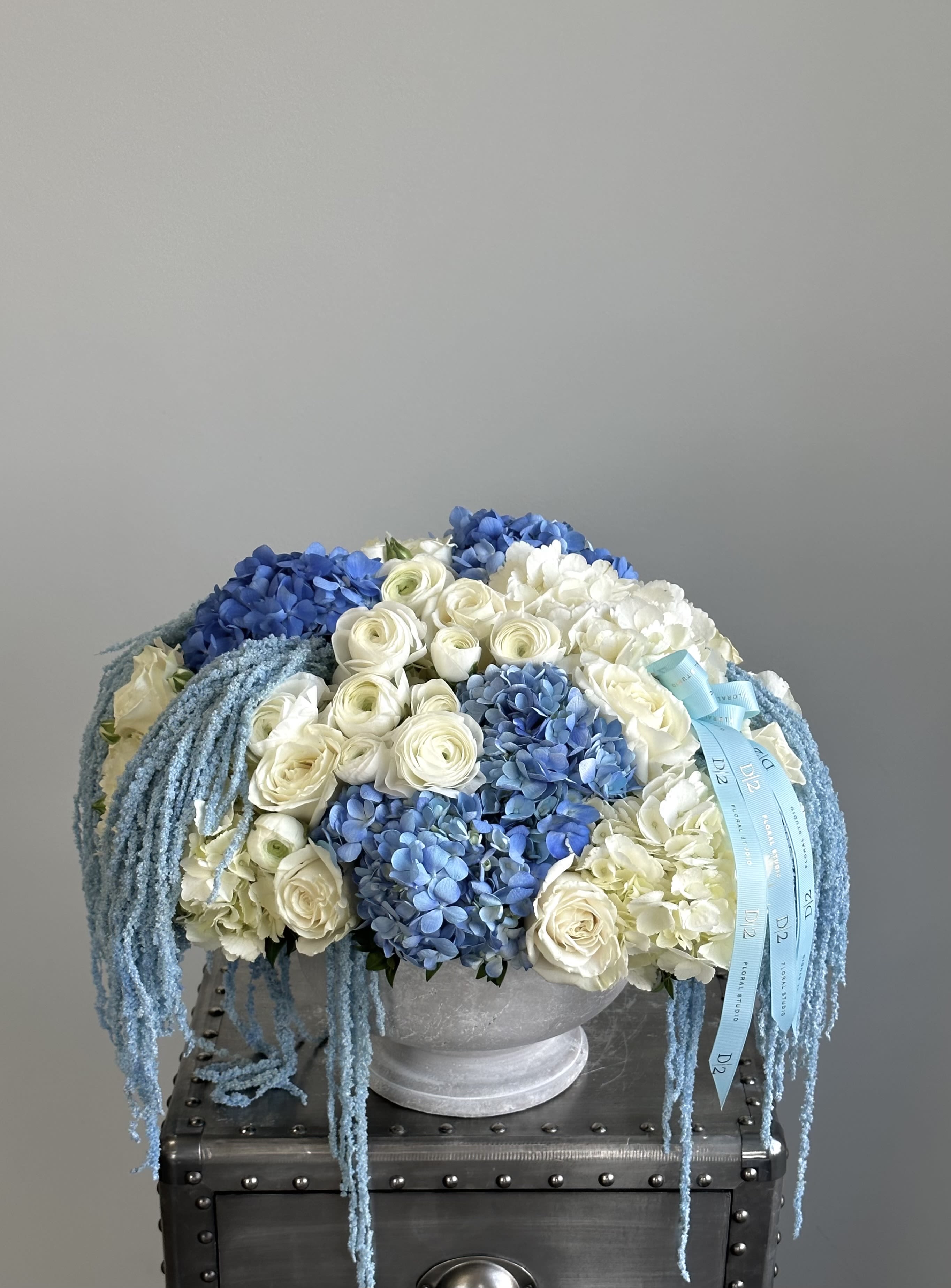 David - This elegant floral arrangement features a lush combination of creamy white roses and ranunculus, paired beautifully with rich blue and light blue hydrangeas. Cascading blue amaranthus adds a soft, flowing texture, while a light blue ribbon branded with “D|2” adds a custom, luxurious touch, set in a classic white urn vase.