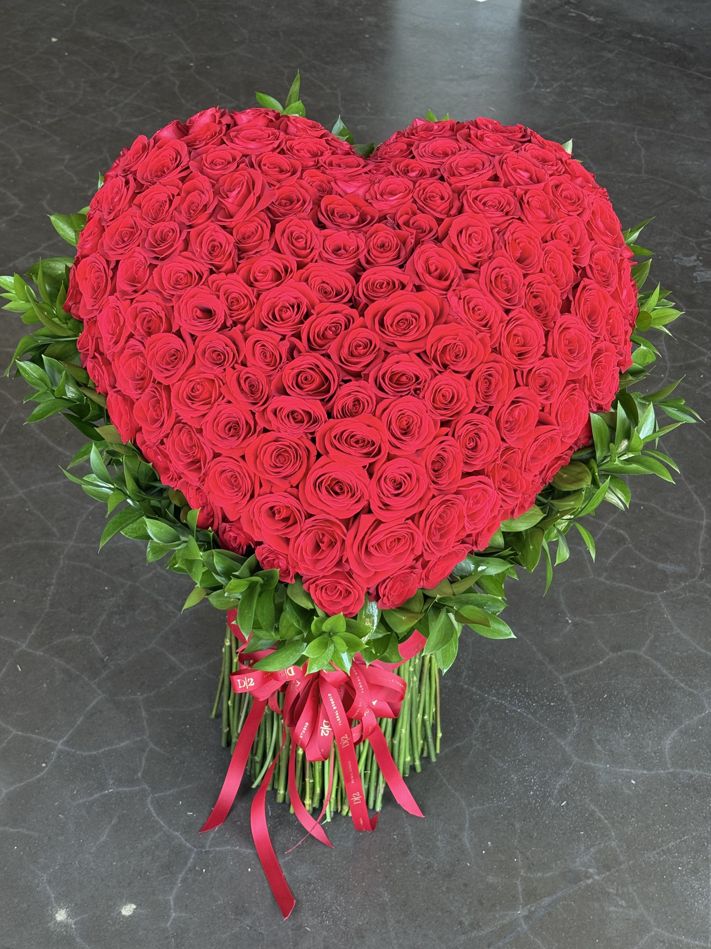 Harmony - A breathtaking heart-shaped arrangement composed of vibrant red roses, elegantly framed with lush greenery and finished with bold red ribbons at the base—an unforgettable expression of deep love and passion, perfect for romantic milestones or grand gestures.