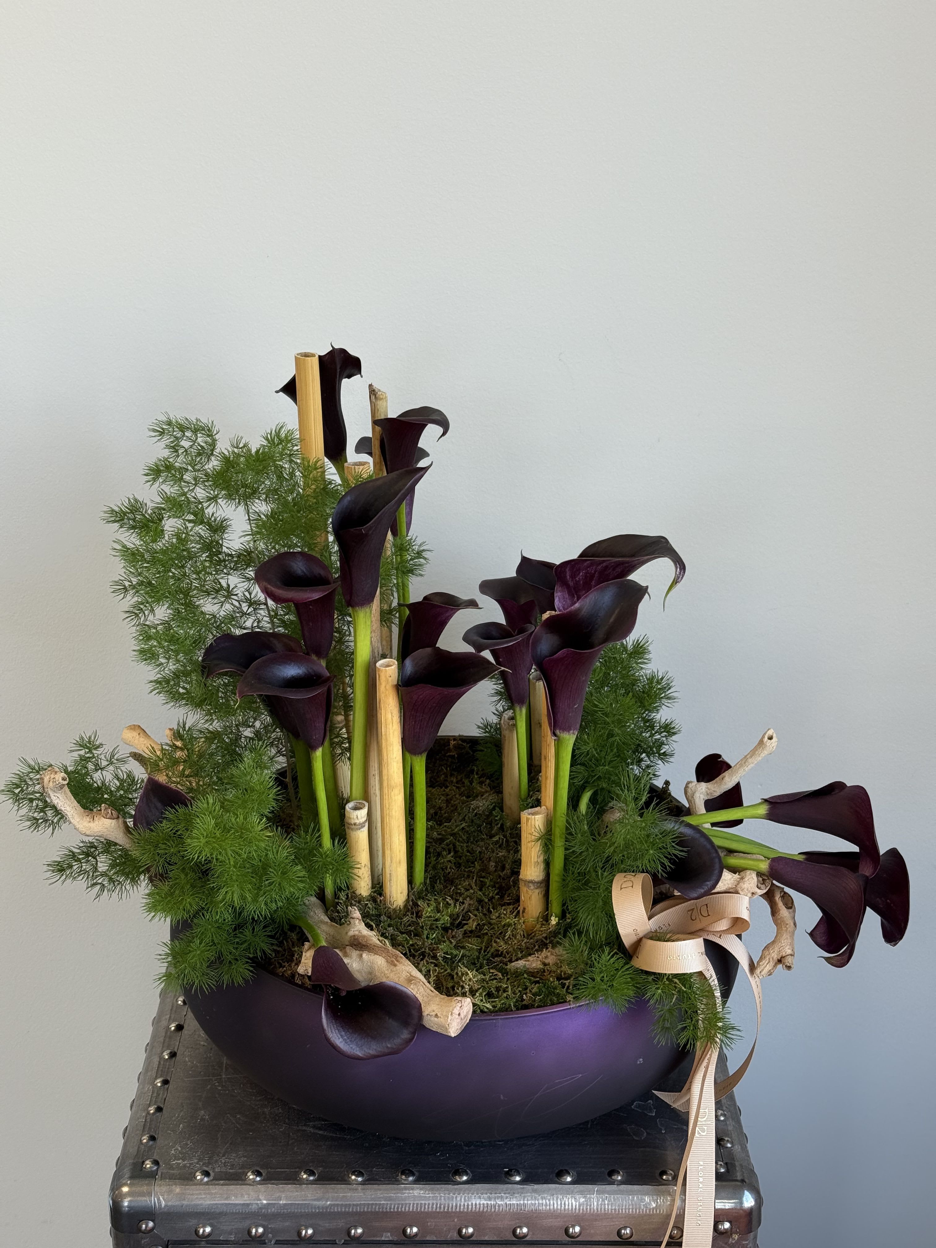 Dark purple calla lilies arranged in a low bowl with greenery and bamboo sticks