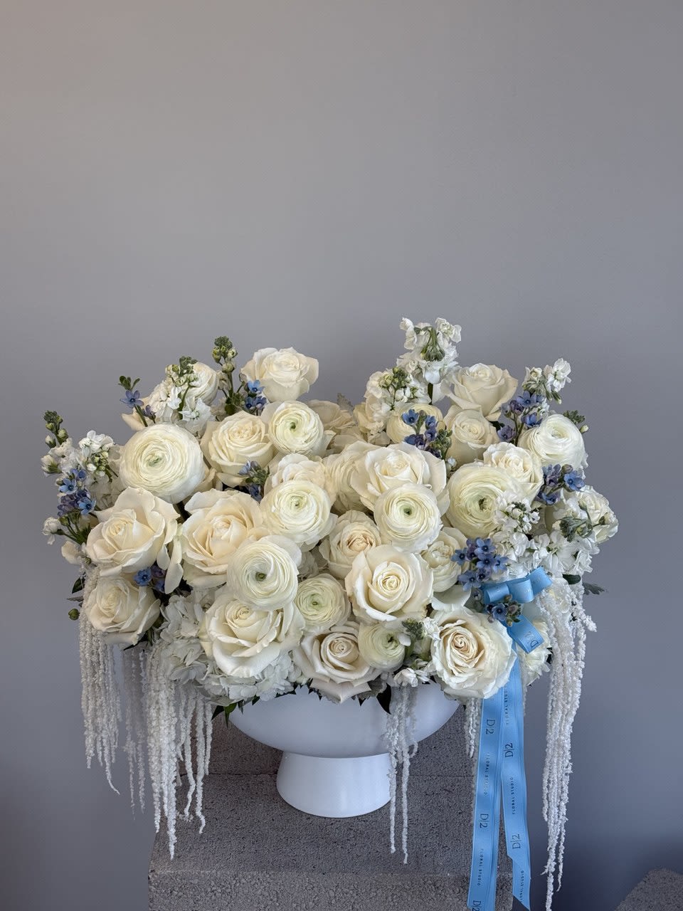 White rose and ranunculus arrangement in a white pedestal vase with blue ribbon