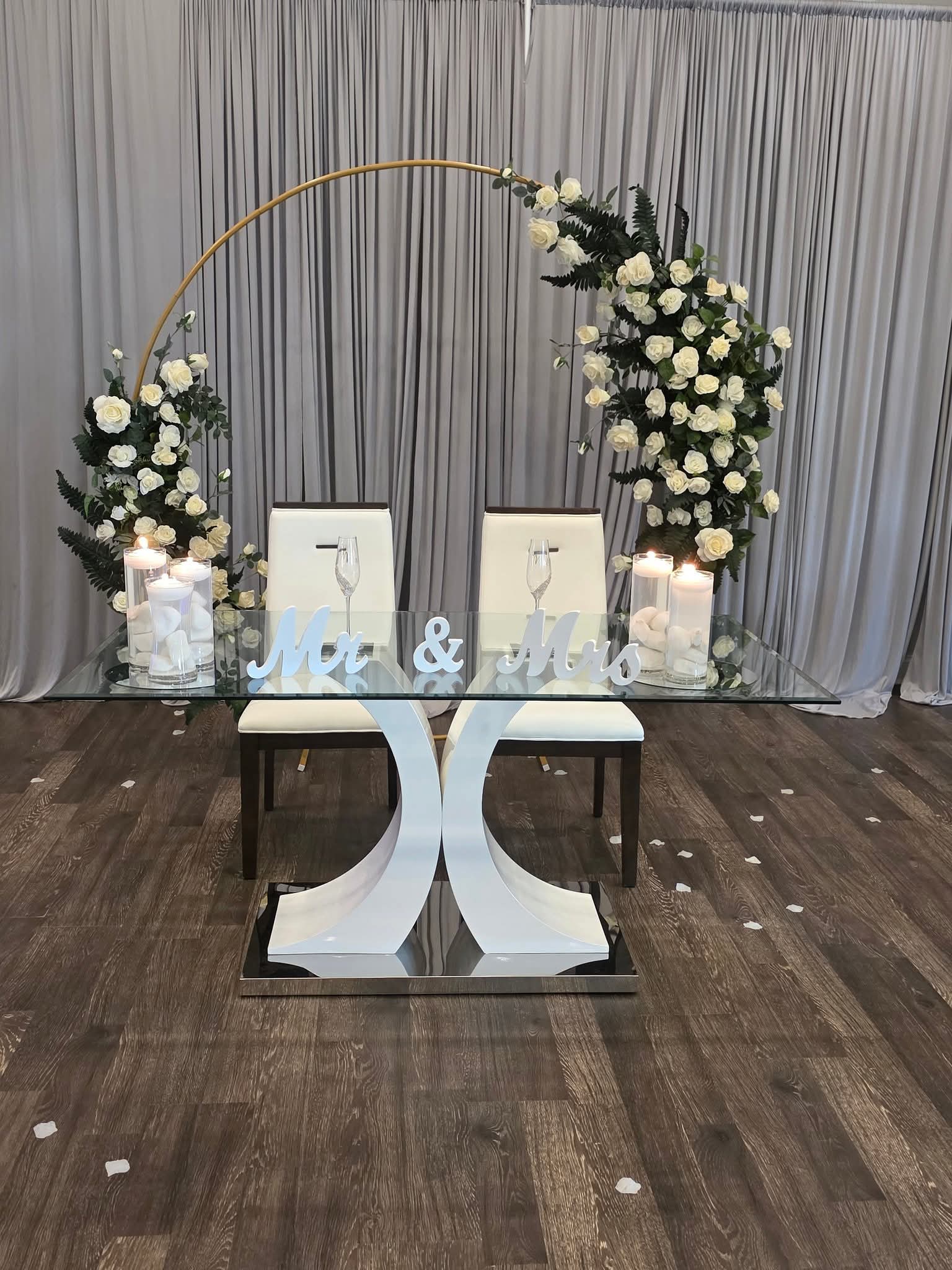 White rose arch over a glass table with candles and "Mr &amp; Mrs" sign
