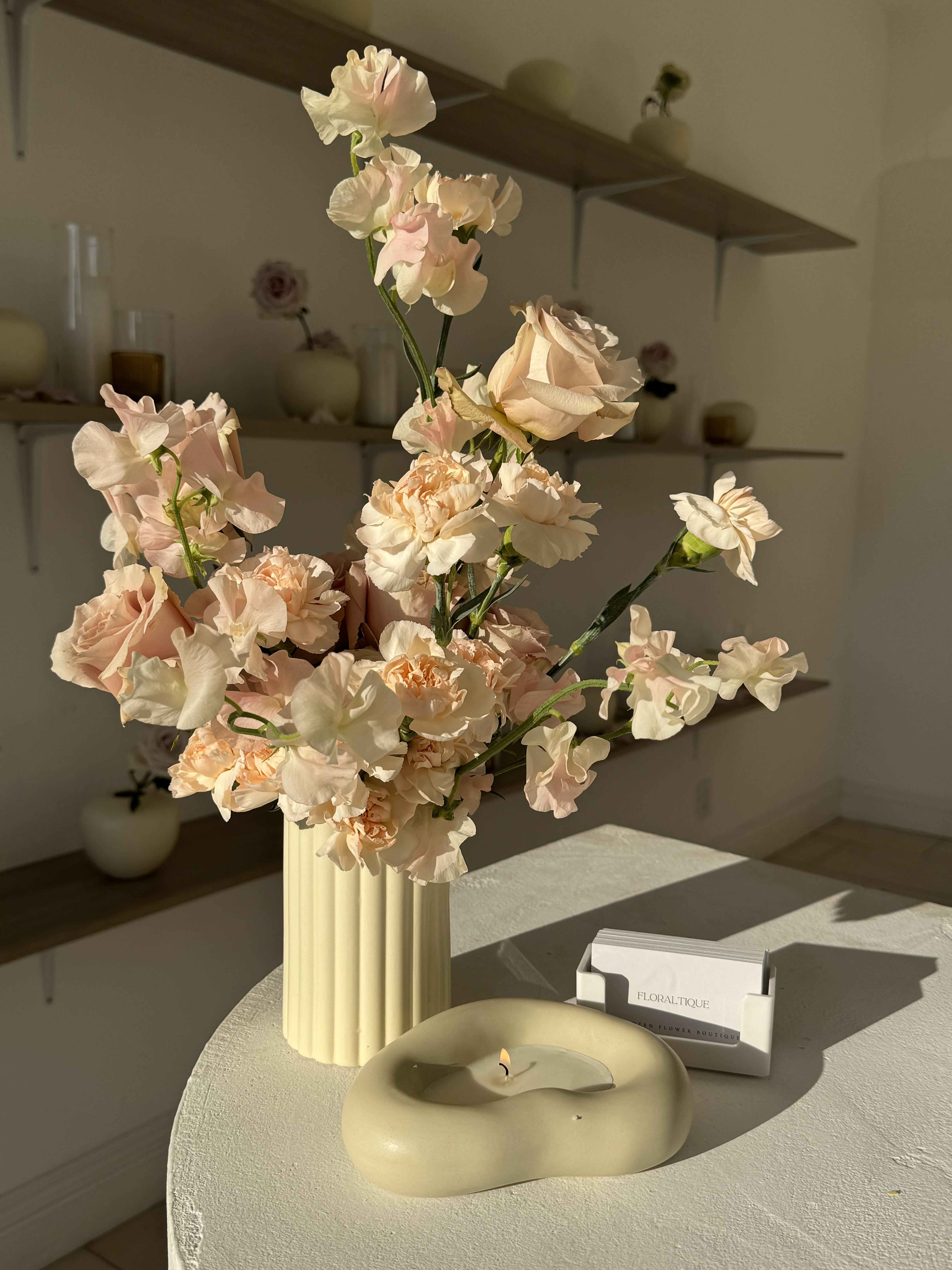 Blush and cream flowers in a ribbed vase beside a candle and card holder