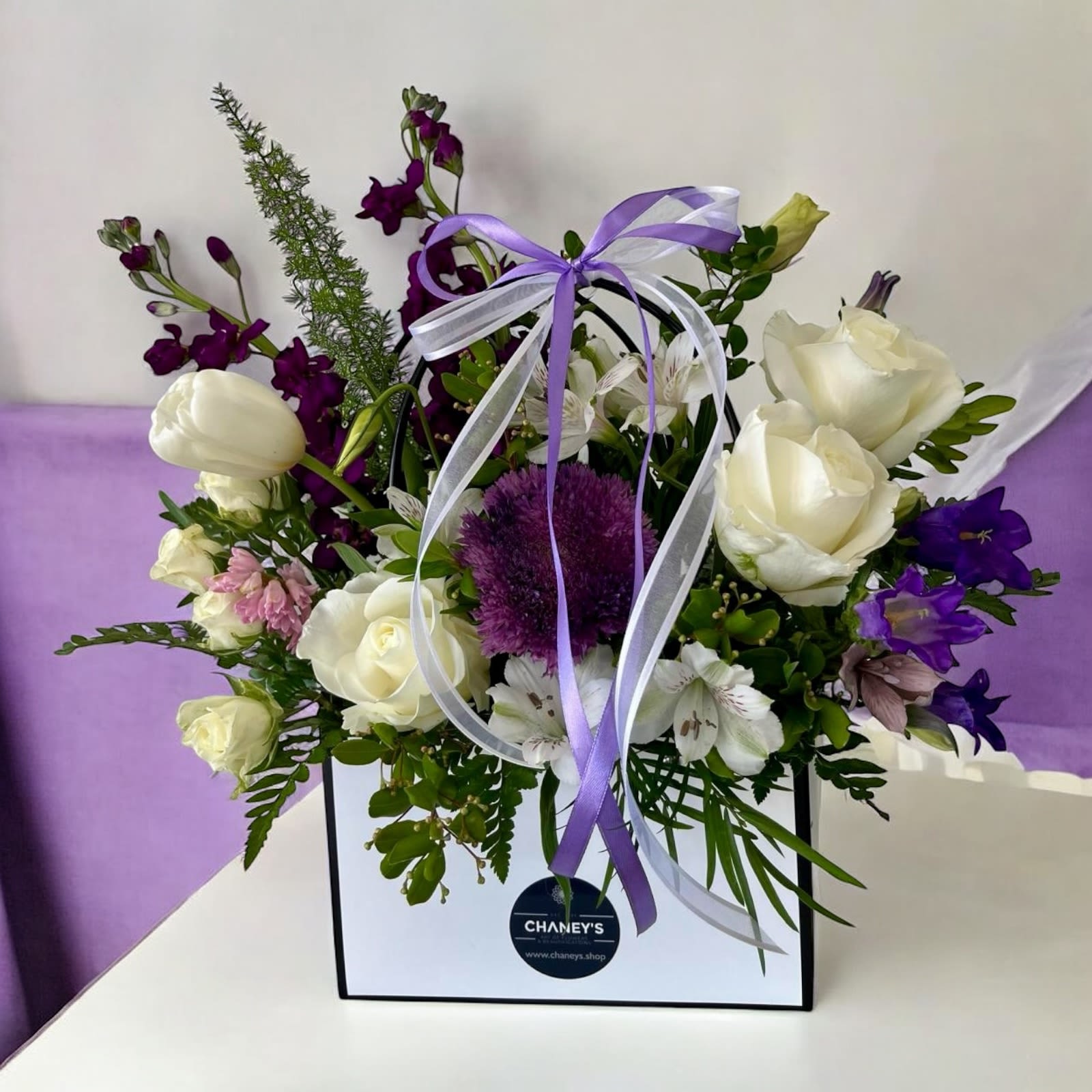 Purple and white floral arrangement in a gift bag with ribbon