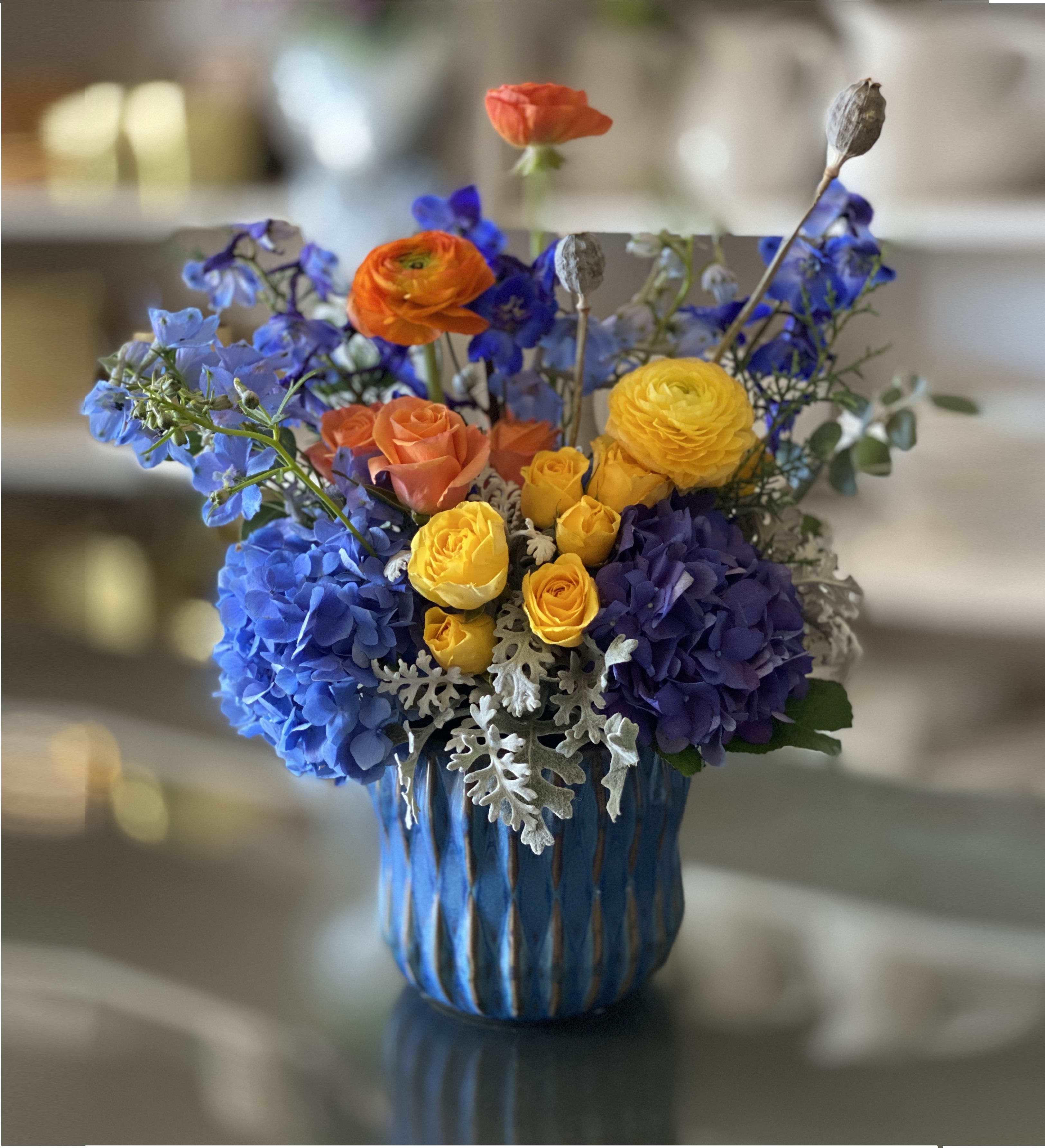 Monet - FB - Introducing Monet – the bold and beautiful bouquet inspired by the captivating paintings of French artist Claude Monet. This eye-catching arrangement is guaranteed to brighten up any room. Sure to be the talk of your next gathering, this exquisite bouquet contains bright blue hydrangeas, sunny yellow and orange ranunculus, and stunning blue delphinium, all creatively arranged in a brilliant blue vase. Whether you're giving it as a present to a loved one or keeping it for yourself, the Monet bouquet will bring a pop of color and cheer into any home. So why not indulge yourself or someone special today with the luxurious Monet bouquet? Brighten up your day with this bold, beautiful masterpiece!  Approximate Arrangement Size 16H X 15W