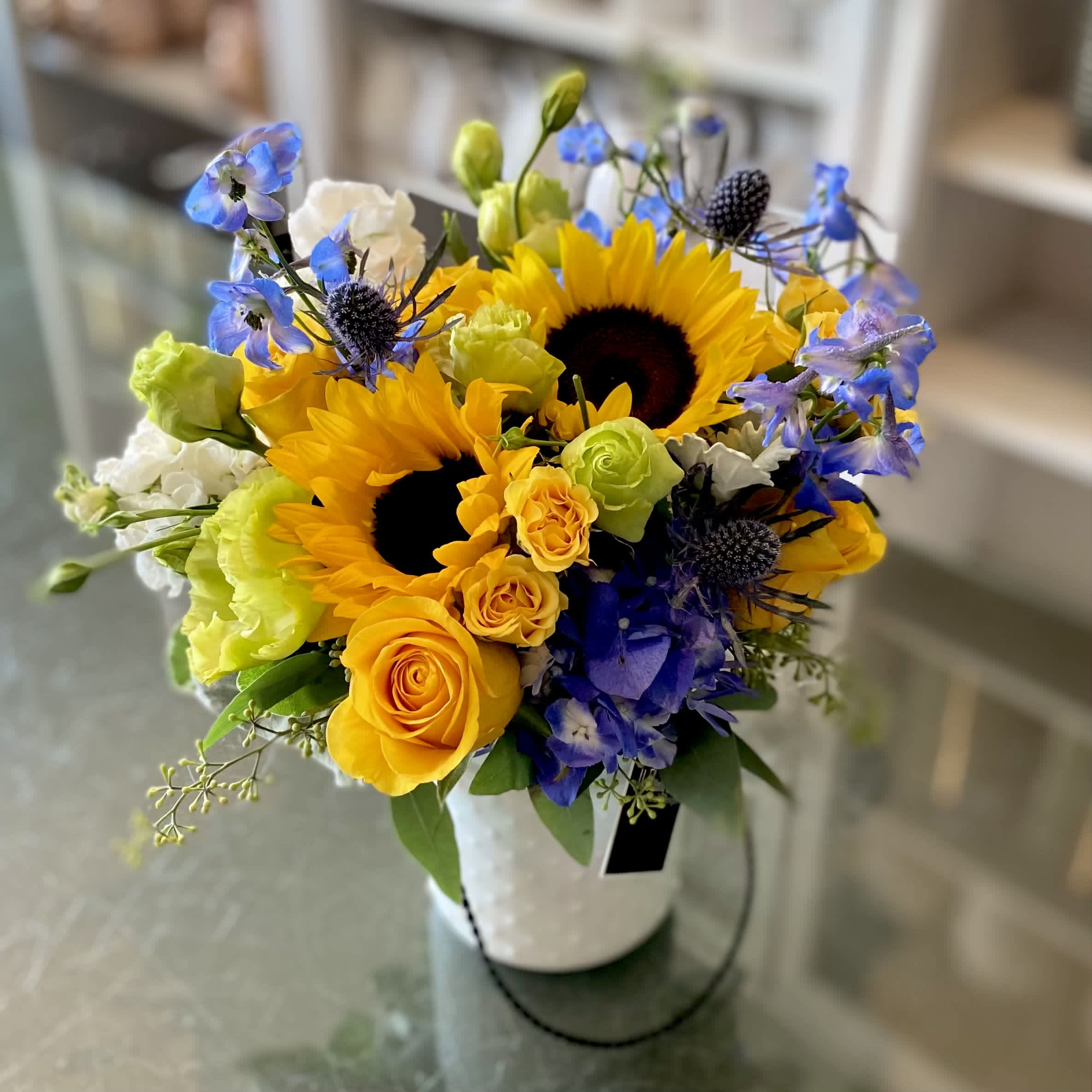 Here Come's The Sunv - FB - Greet the day with timeless joy with Here Comes the Sun – an arrangement inspired by the iconic Beatles song of the same name. This rustic vase, filled with a cheerful bouquet of flowers in shades of sunny yellow and bright blue will have your family and friends feeling brighter since. A burst of yellows make this perfect for any special occasion; from a summery event to a ‘just because’ gift. Yellow roses are surrounded by sunflowers, hydrangeas, lisianthus, thistle and blue delphinium – bring sunshine into any room without having to step foot outside. The Beatles lyrics come to life with Here Comes the Sun, a song full of hope that is certain to bring exactly that to your day!
