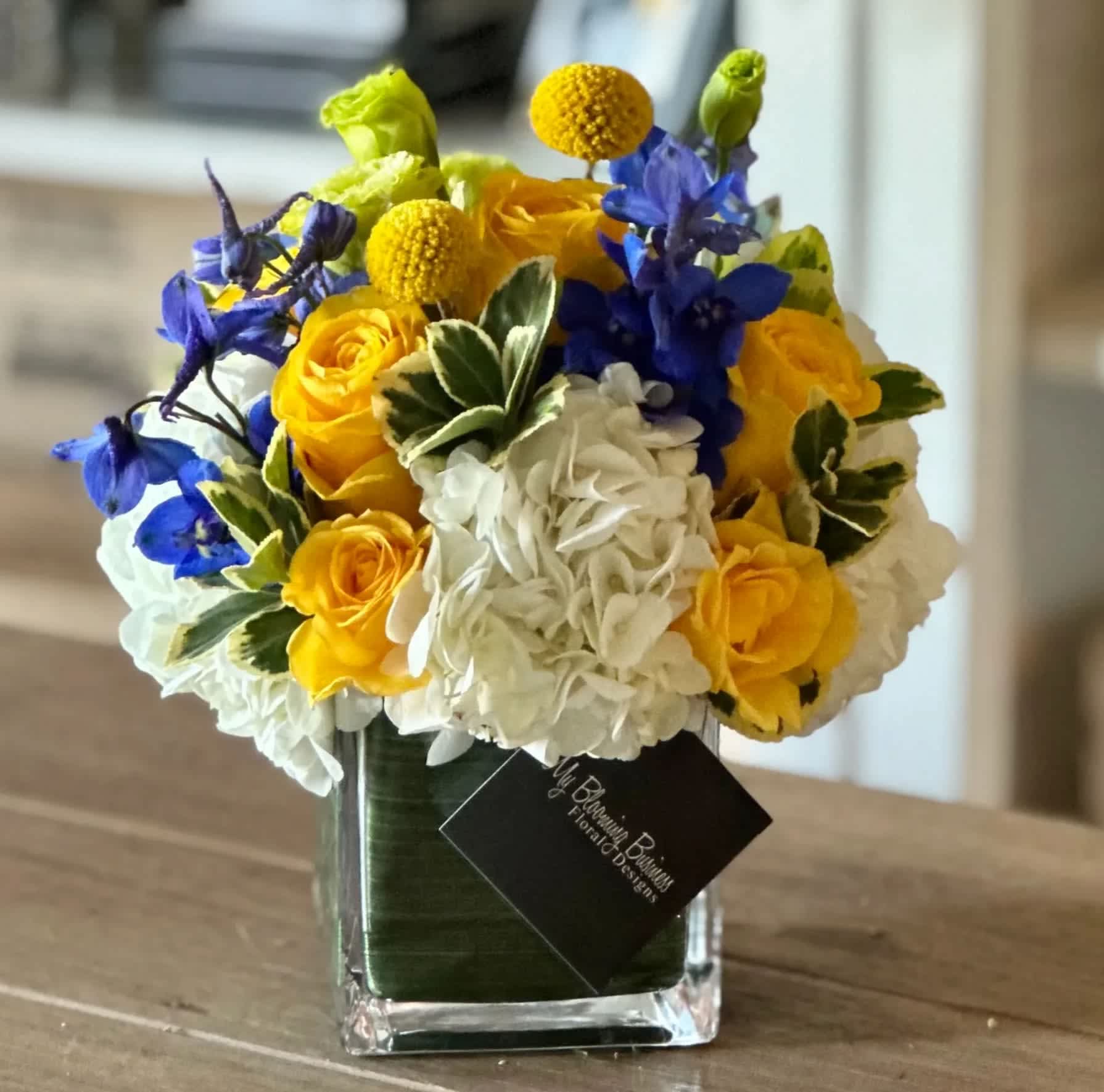 Bluebell - FB - 4X4 glass cube arrangement.  Our bluebell arrangement is a mix of yellow florals with a hint of blue florals  in a clear cube vase with green t leaf lining inside. the perfect arrangment for any occasion to send to your love ones. 
