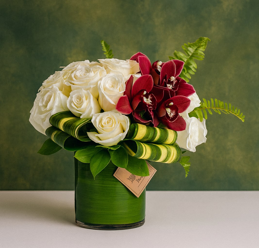 Mischa - FB - Introducing Mischa – the perfect flower arrangement to bring warmth, elegance, and joy into any space! This gorgeous ensemble of newly cut white vandela roses and burgundy cymbidium orchids is housed in a trendy and clear cylinder vase lined with a beautiful T leaf pattern. Mischa truly encapsulates the beauty of nature inside your home, allowing you to enjoy the vibrant colors all year round. 