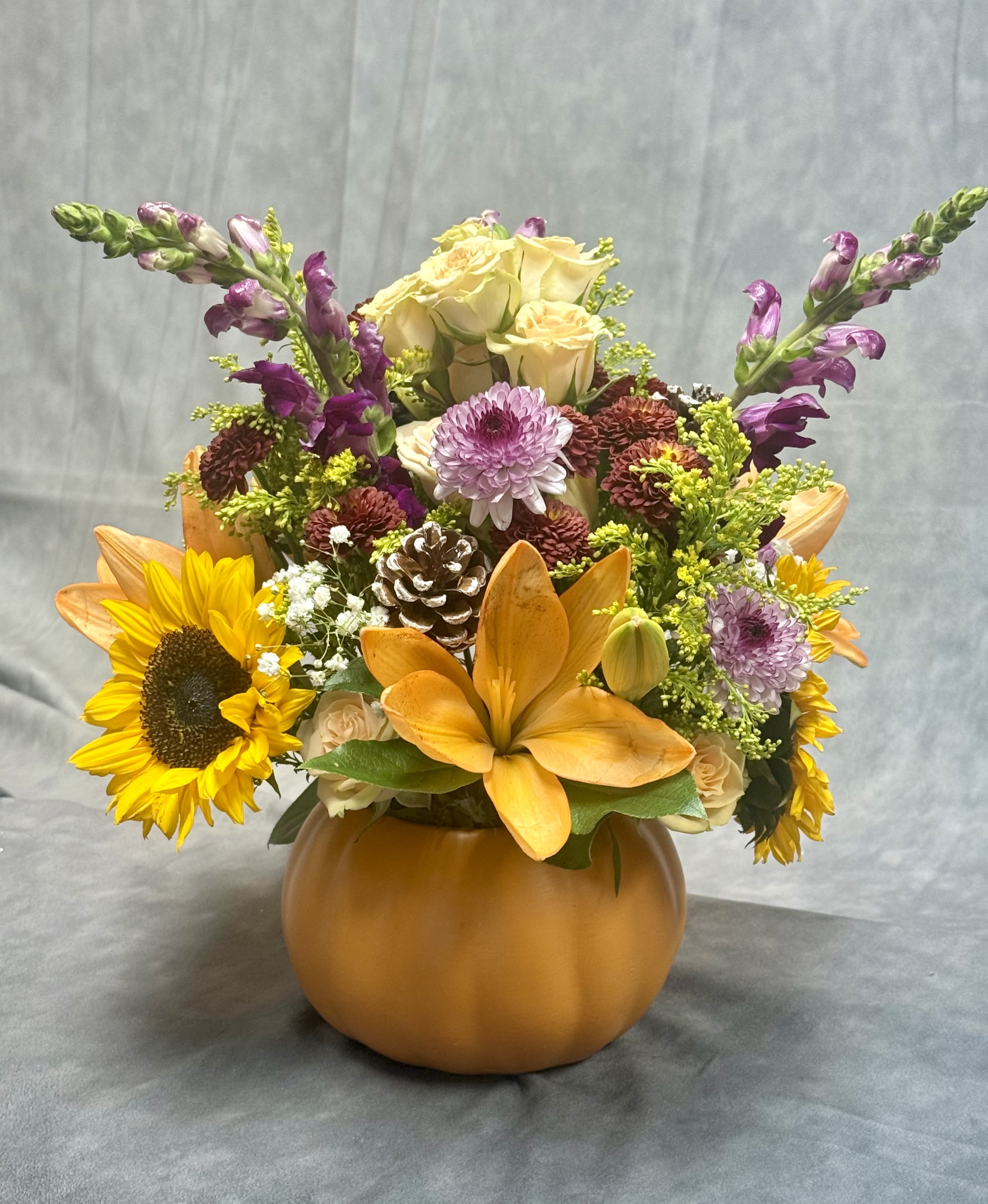 Pumpkin Spice Blooms - Bring fall into your home with this vibrant pumpkin arrangement filled with golden sunflowers, orange lilies, soft roses, colorful mums, snapdragons, and rustic pinecones.