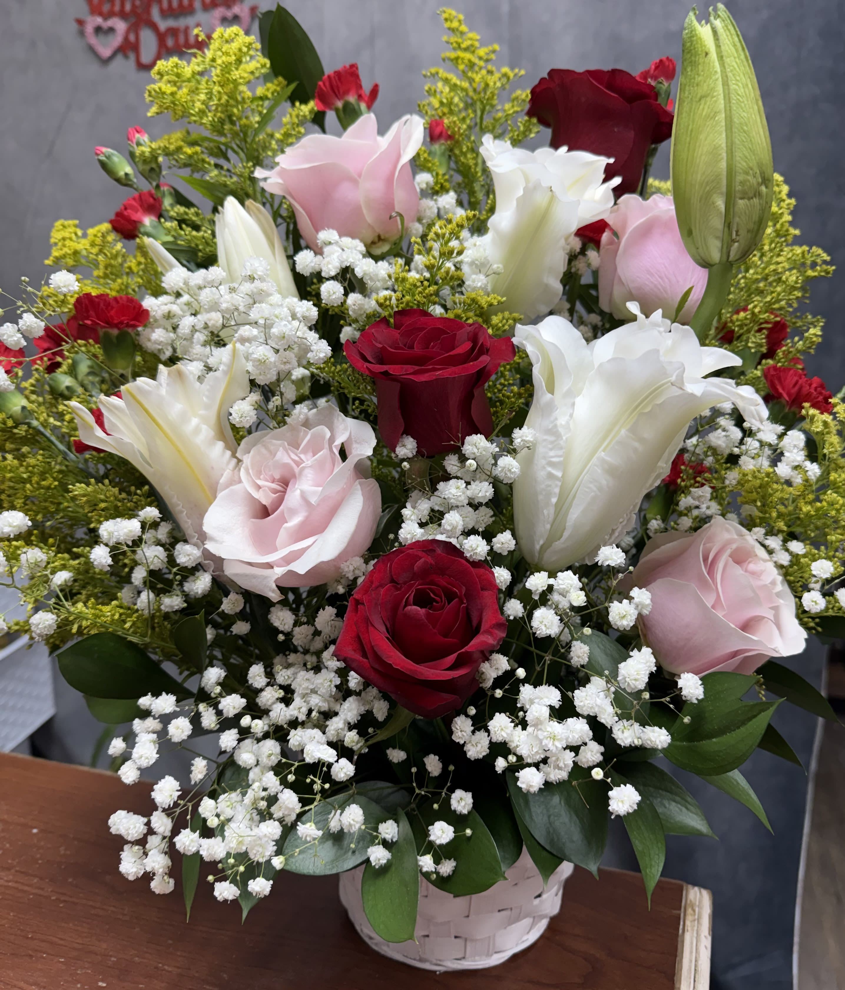 With love basket - Red, pink and white roses and lilies with red carnations, baby's breath and yellow fillers in a white woven basket-warm, romantic arrangement for birthdays, anniversaries, or thoughtful gestures.