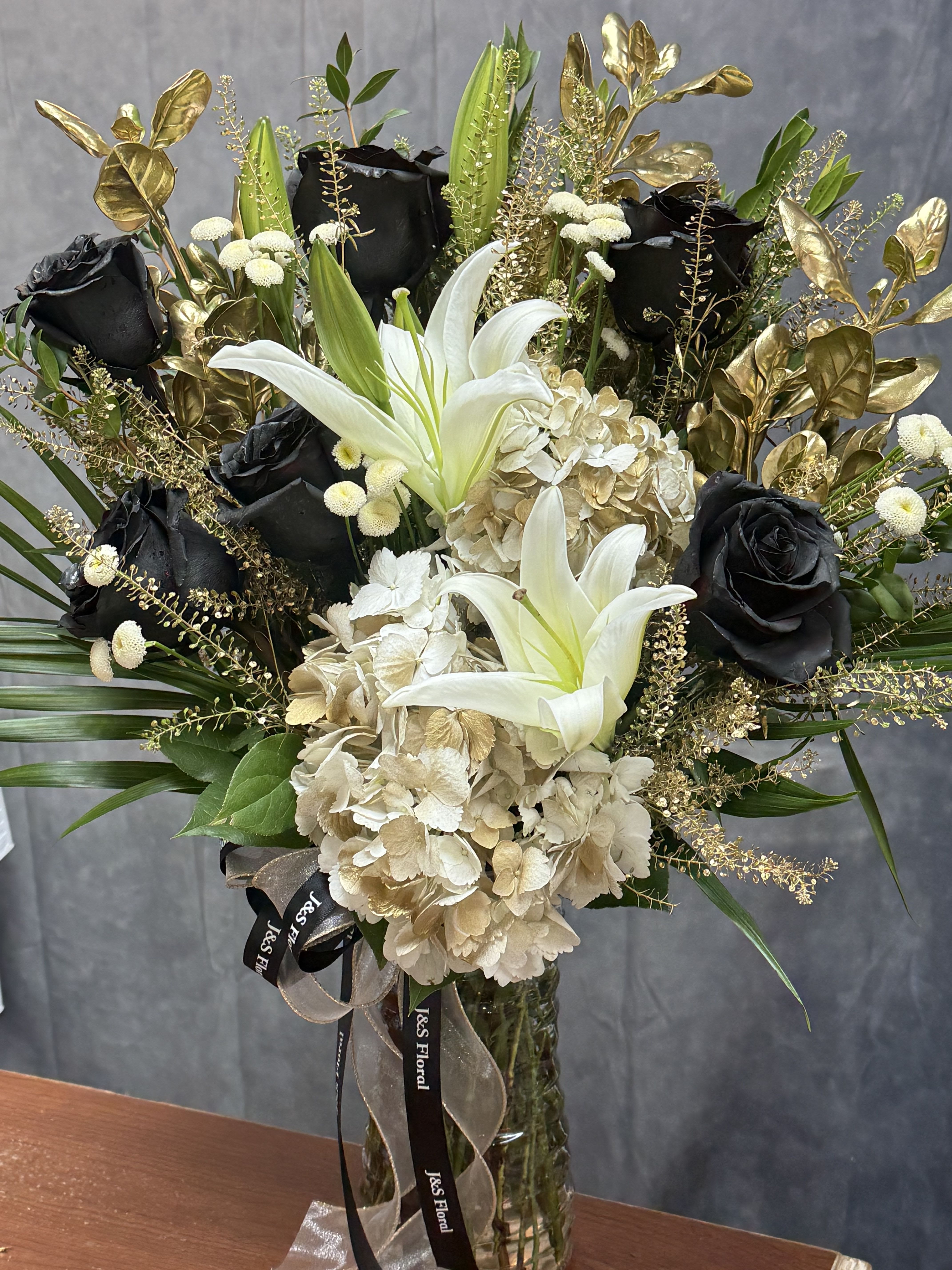 Midnight Gold Vase - White lilies, black roses and cream gold hydrangeas with metallic gold foliage in a glass vase. Dramatic, high-contrast arrangement ideal for elegant anniversaries, or sophisticated celebrations.