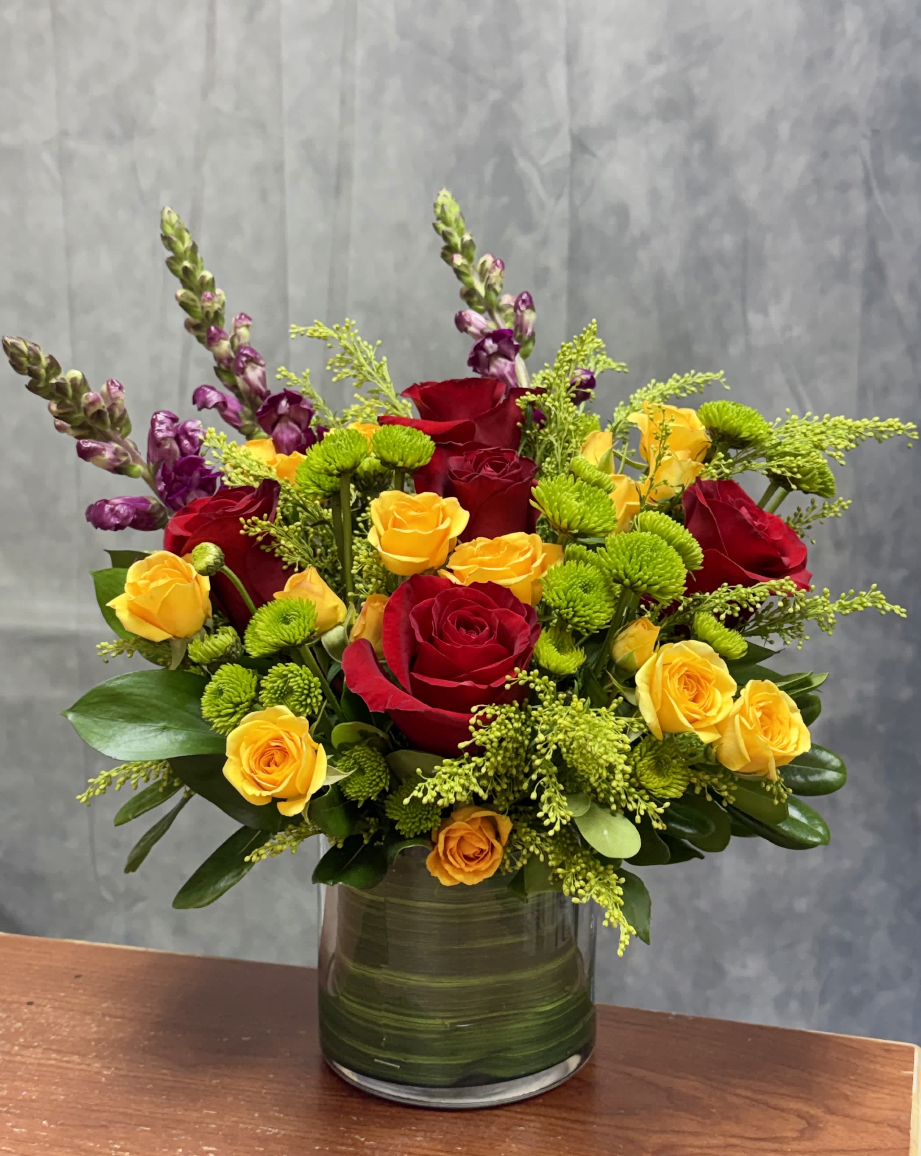 Red and yellow roses arranged in a glass vase with purple spikes