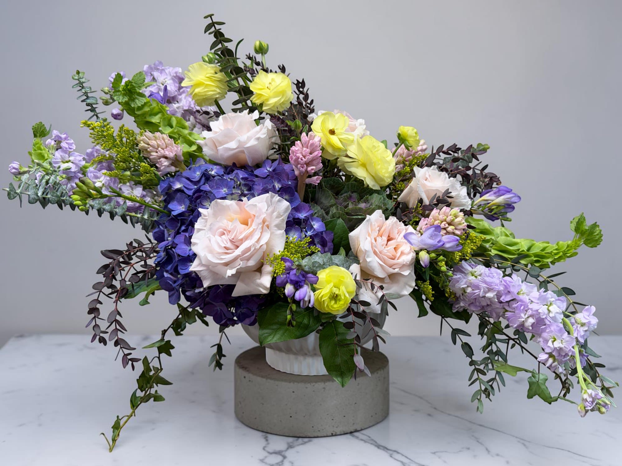 WCF Luxe Collection - Designer's Choice Customizable Compote Arrangement - The Designer's Choice Compote Arrangement is an artistic and elegant arrangement of the highest quality blooms arranged by our top floral designers. Perfect for a grand display on a dining table or in a foyer. The standard arrangement typically contains the following flowers based on availability: premium hydrangeas, stock, roses, ranunculus or anemone, freesia, and unique seasonal blooms with flowing greenery in fabulously designed color palettes. The deluxe version contains the standard flowers plus lisianthus. The premium version contains the standard flowers plus lisianthus and dahlias.  Do you have favorite flowers that you want included? Choose your own favorite colors and blooms at checkout by listing them in the special instructions section. Delivered in a sophisticated white ceramic compote planter. Approximately 36" W x 20" H  Please note that orders must be placed a least 48 hours prior to fulfillment date in order to procure the freshest and highest quality blooms. 