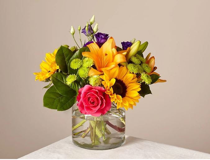 Mixed bouquet in a clear glass vase with a pink rose, sunflower, lily, and purple blooms