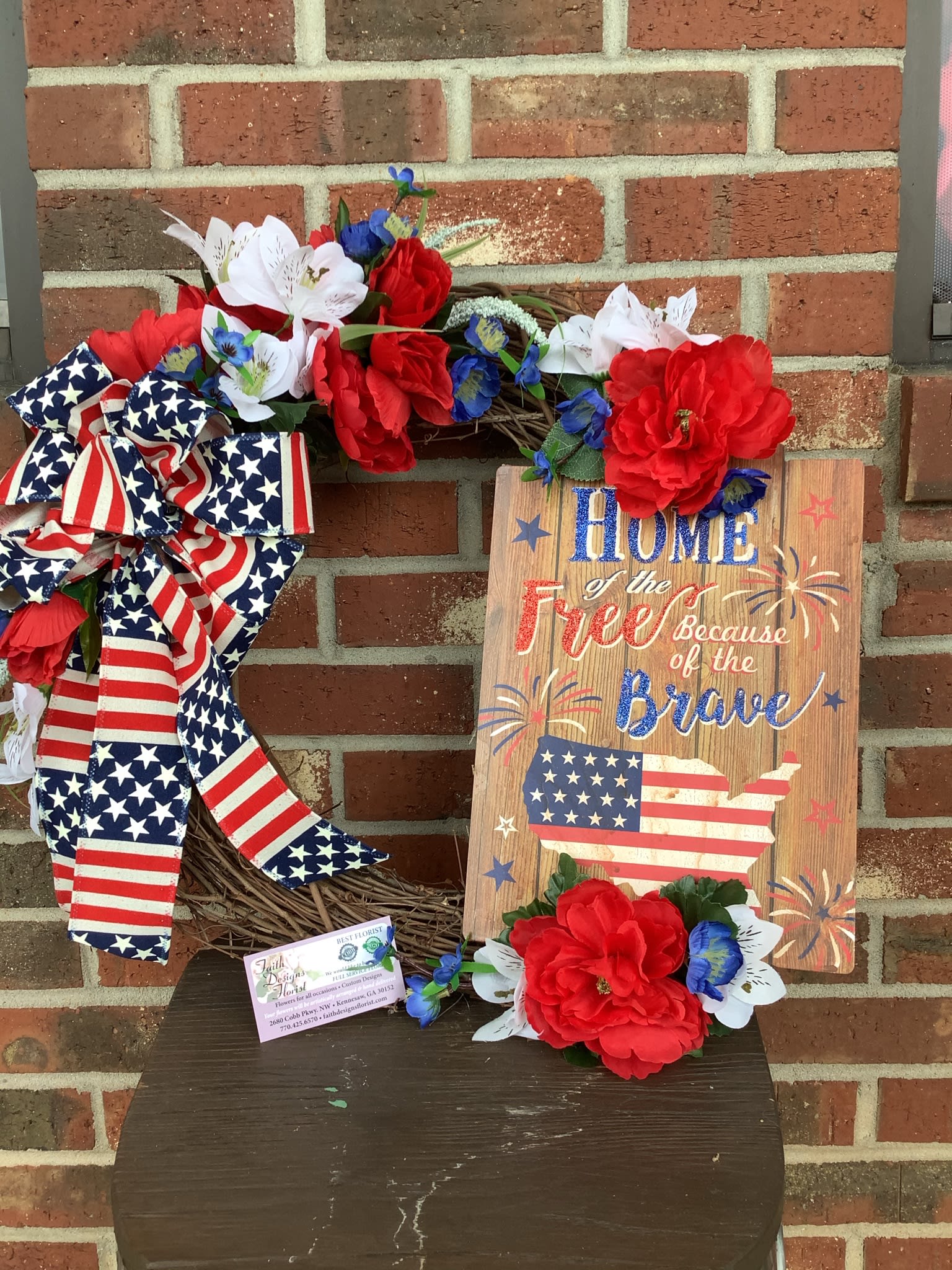 Patriotic wreath with red, white, and blue flowers and a flag ribbon beside a wooden sign
