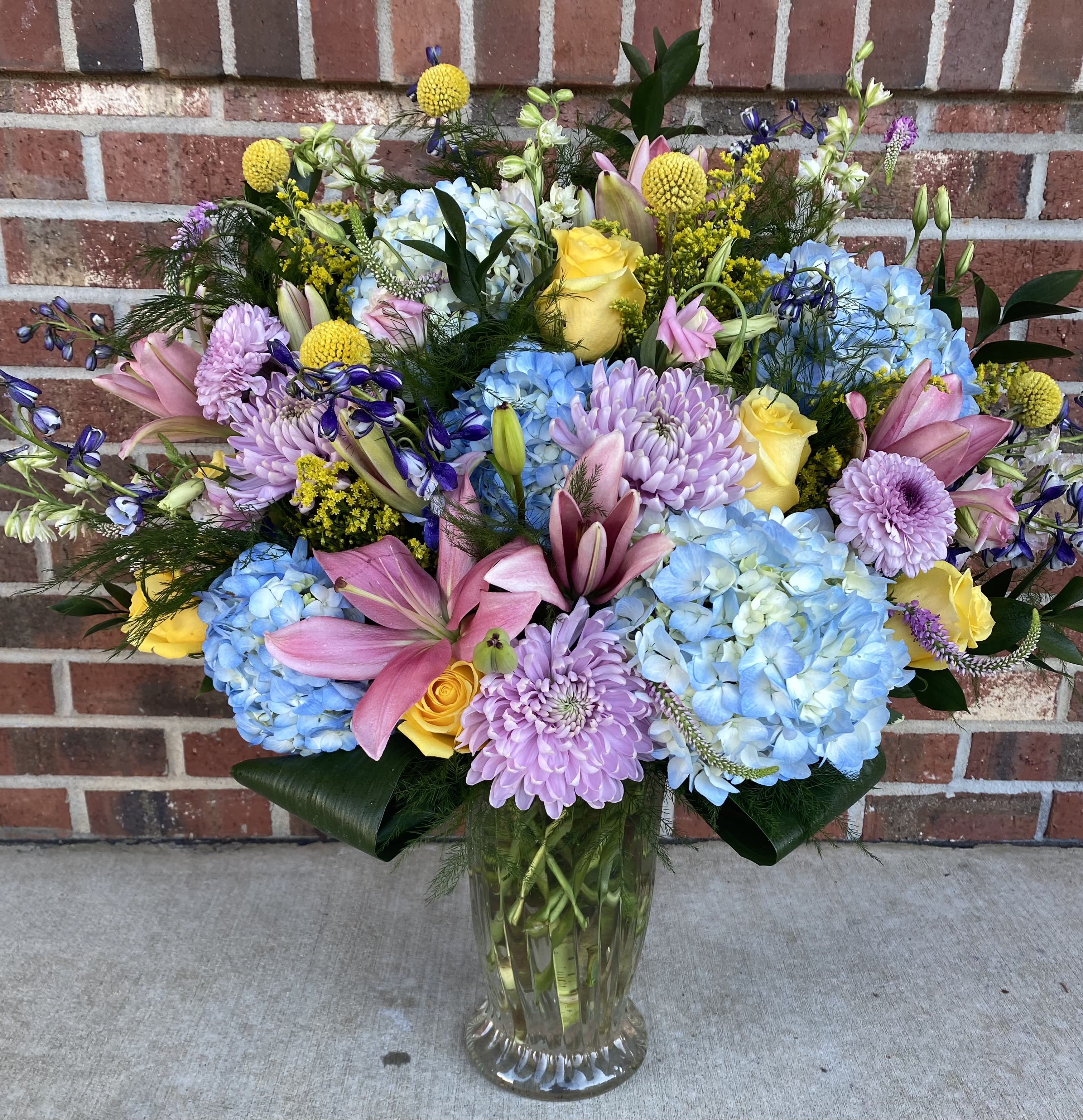 Mixed bouquet of blue hydrangeas, pink lilies, and yellow roses in a glass vase