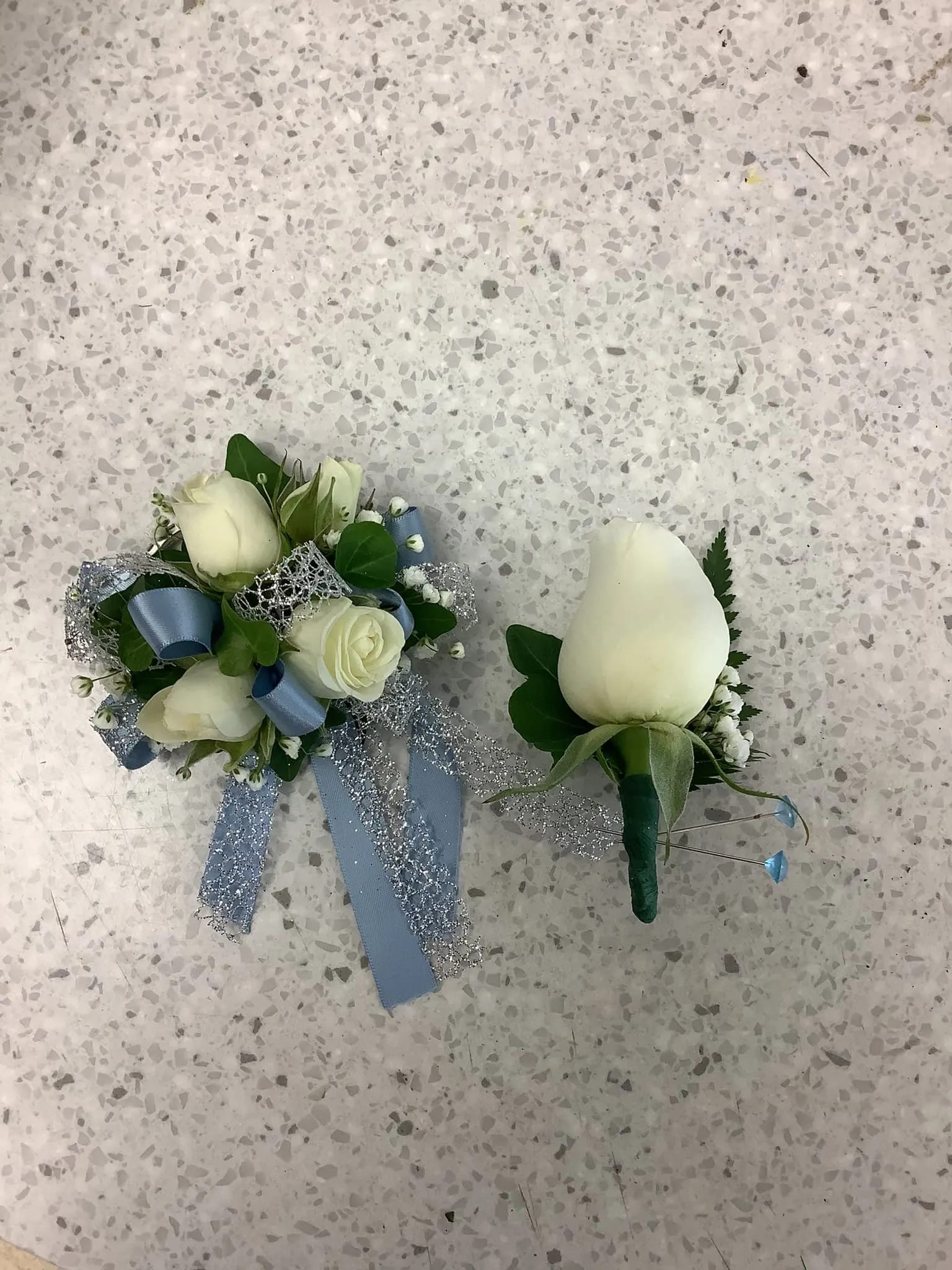 Two white rose boutonnieres with blue ribbon accents on a speckled floor