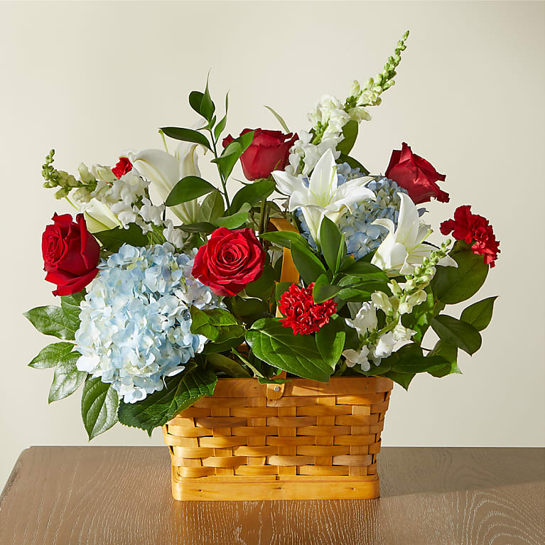 Greater Glory Basket - A full, rounded tabletop arrangement that pairs bold red roses and carnations with pristine white lilies and snapdragons-perfect as a Mother's Day gift or a spring centerpiece. The Greater Glory Basket's red, white, and blue palette also lends a respectful, patriotic tone for national holidays or remembrance while remaining warm and celebratory for birthdays or get-well wishes. The overall impression is one of thoughtful dignity that will make the person you're thinking of feel honored and cared for.  This design combines soft blue hydrangea clusters for a cool accent with lush greenery tucked among the blooms, all gathered in a warm-toned woven wood basket. Flowers rise slightly above the rim for a medium, tabletop presence that's easy to display on a mantel, dining table, or hospital bedside. Care is simple: keep the floral foam hydrated and place away from direct heat or drafts to extend freshness.
