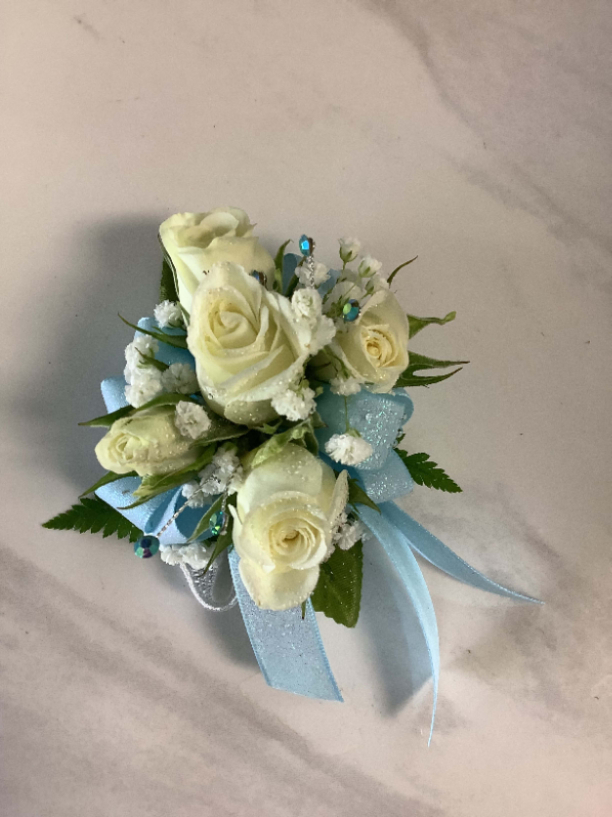  Blue Shimmer Corsage - Creamy white roses gathered in a compact wrist corsage give a quietly elegant first impression-perfect as a birthday gift or to mark prom, graduation, or a wedding celebration. The arrangement's blue and white color story reads fresh and celebratory, so it also makes a thoughtful "thinking of you" gesture or a gentle congratulations for a new baby.  Tightly clustered spray roses and tiny white filler blossoms form the floral center, surrounded by soft loops and tails of light blue and silver ribbon for movement and polish. Small blue jeweled picks tucked among the blooms add a subtle sparkle and a formal touch without overpowering the delicate palette. The compact size sits comfortably on the wrist and coordinates easily with gowns, graduation robes, or nursing-visit outfits.  This corsage is designed to make the recipient feel noticed and cherished on special days-from prom night to a mother's first Mother's Day or a proud graduation. Order with confidence for same-day pickup or local delivery when available; to keep it looking its best, store cool until worn and mist lightly if needed.