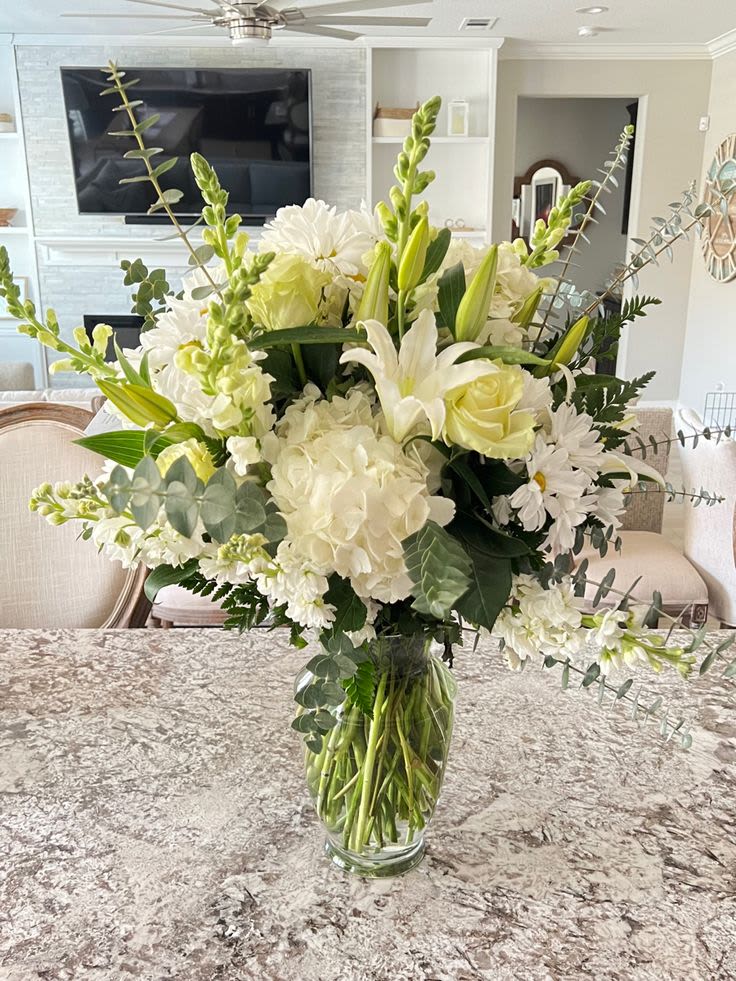 Peaceful Passage - A soft, airy bouquet of white and pale green blooms that feels quietly comforting - perfect as a sympathy arrangement or a calming gift for someone who needs a gentle reminder you care. This flower arrangement blends hydrangeas, open lilies, roses, daisy-like blooms, and tall snapdragons for a graceful silhouette that reads both elegant and approachable. The neutral palette and silvery eucalyptus-like accents give it a softly romantic, restful presence ideal for memorials, get-well wishes, or a serene home display.  Arranged in a clear glass vase, the design showcases rounded hydrangea clusters at the base with lily petals and roses interspersed for texture, while snapdragon spikes add vertical movement and visual interest. Pale greens and mixed neutral tones keep the look peaceful and versatile, so it complements bedroom, living room, or bedside settings without overwhelming the space. The glass vase gives a clean finish and makes the arrangement easy to place on a mantle, table, or reception area.  Choose this arrangement when you want the recipient to feel seen and soothed - a meaningful way to say you're thinking of them or to honor a life. For best longevity, keep water fresh and trim stems slightly every few days; the clear vase also makes it simple to monitor water level and refresh as needed.