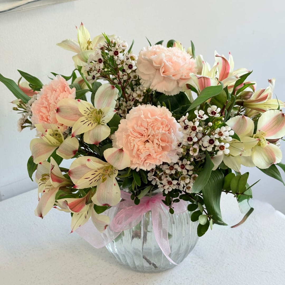 Peach carnations and pale lilies in a clear glass vase with a pink ribbon