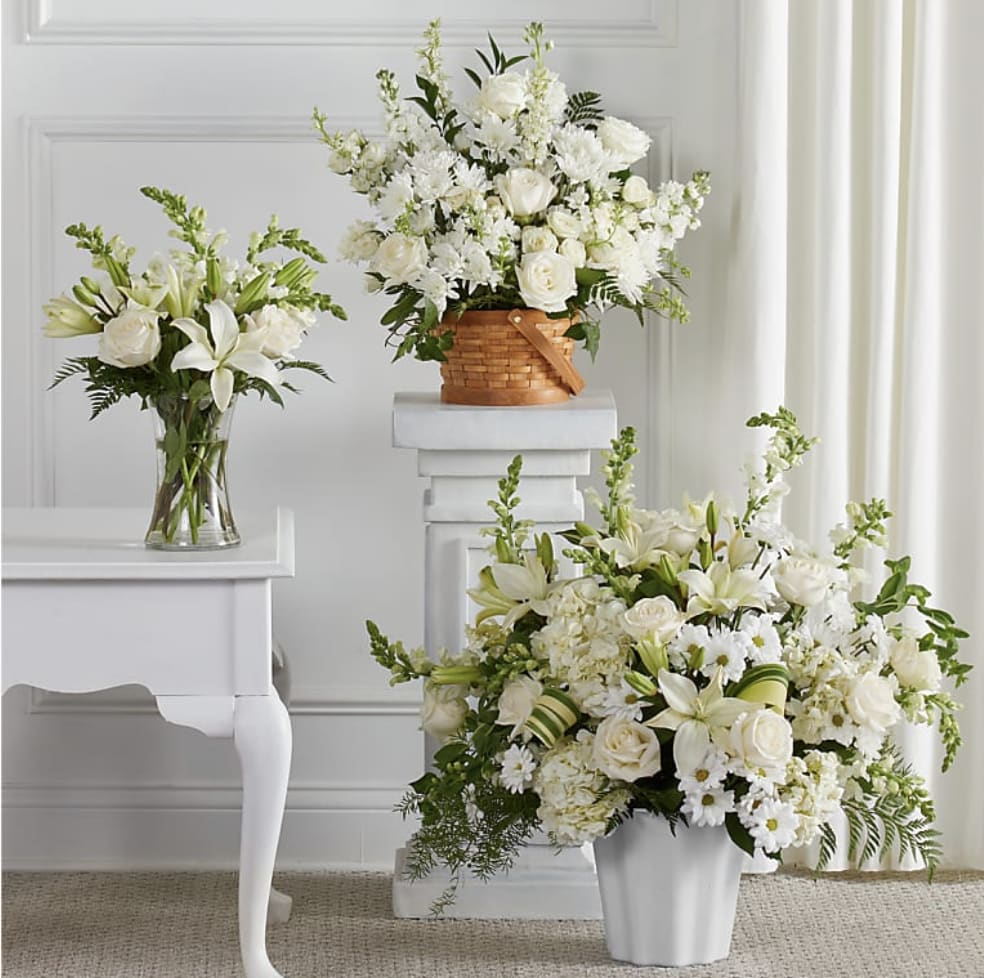 Set of three white floral arrangements with lilies and roses in glass vase, basket, and white container.