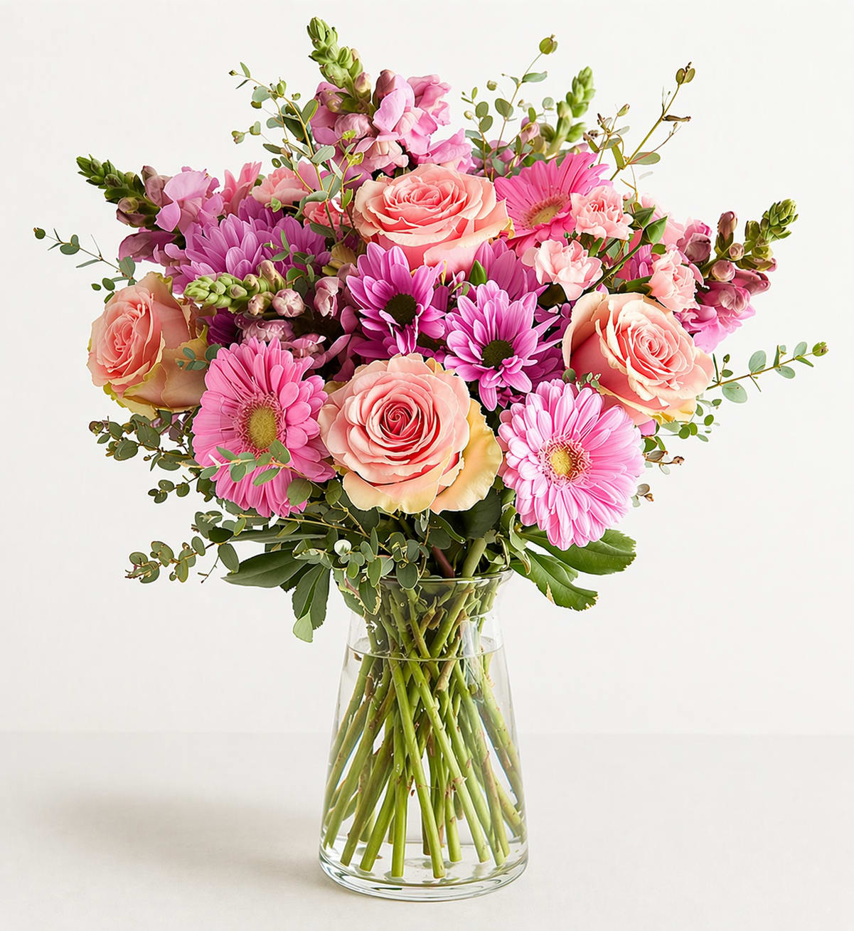 Rosewater - A lush arrangement in a clear glass vase featuring premium roses, gerbera daisies, and seasonal accent blooms in soft pinks and blush tones, finished with fresh greenery. Designed in a full, rounded style with visible stems for a clean, natural look. Flower varieties may vary based on availability while maintaining the same color palette and overall feel.