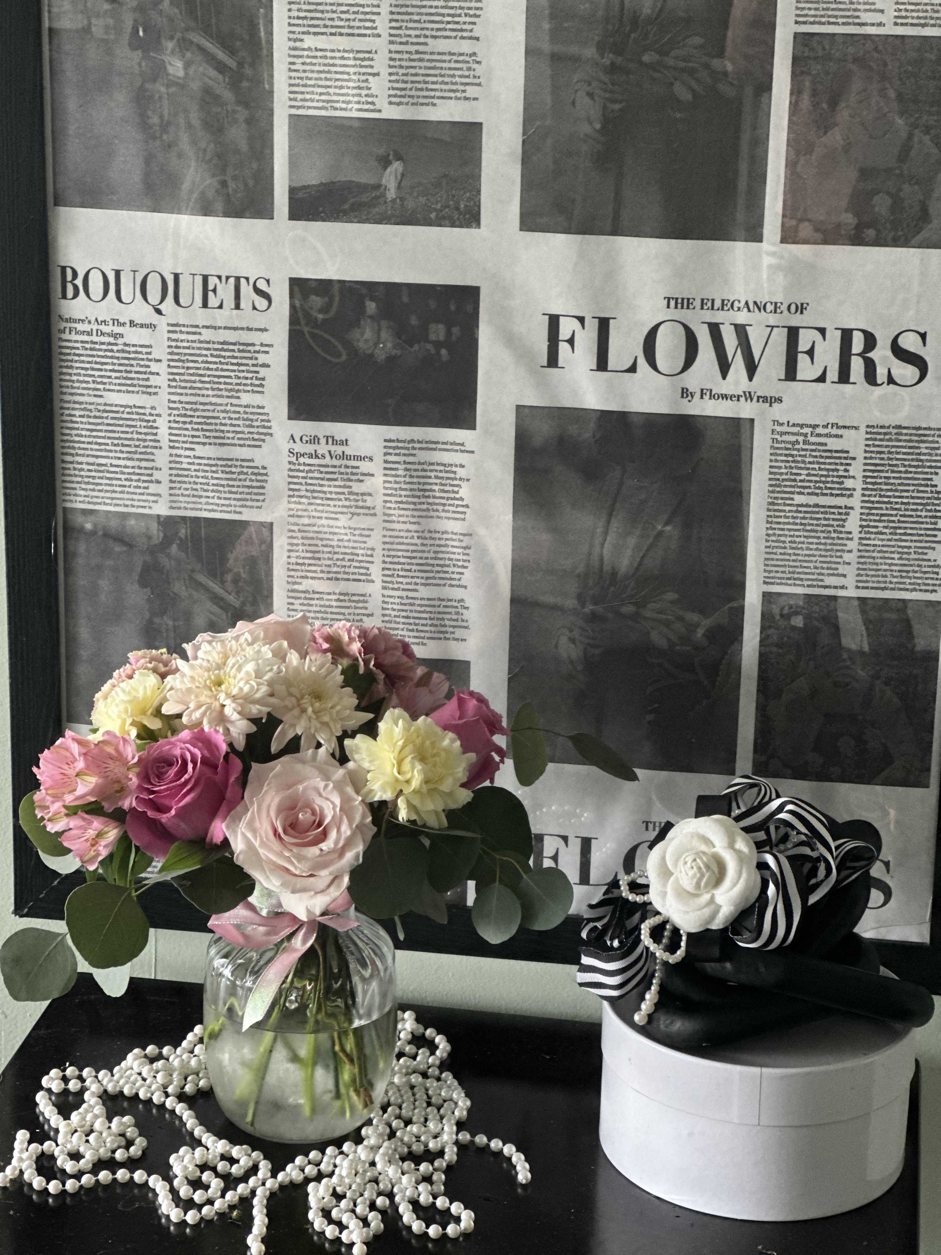 Pink and white flower bouquet in a glass vase beside a small gift box with ribbon.