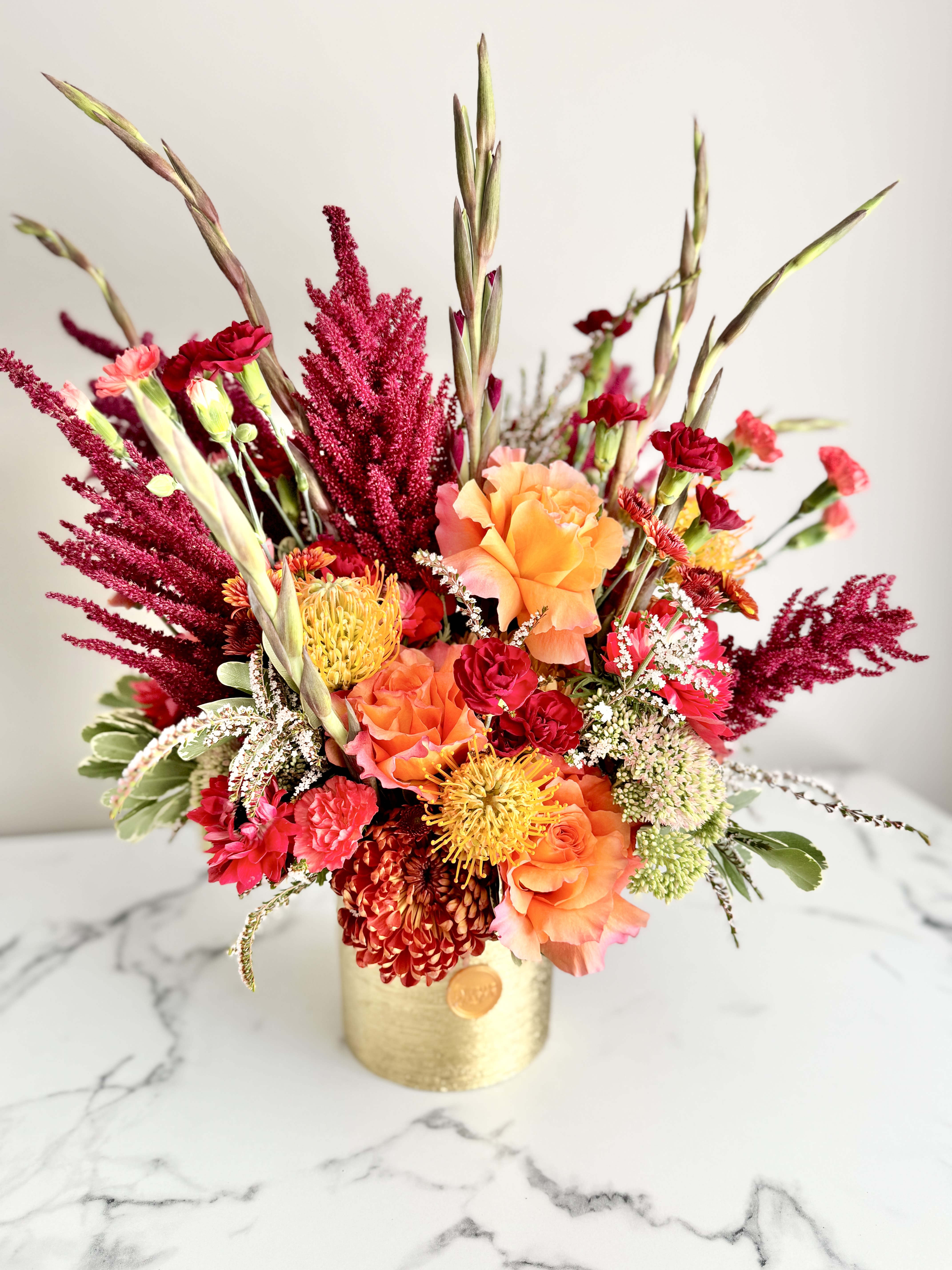 Vibrant arrangement of orange roses, red carnations and yellow protea in a gold cylinder vase