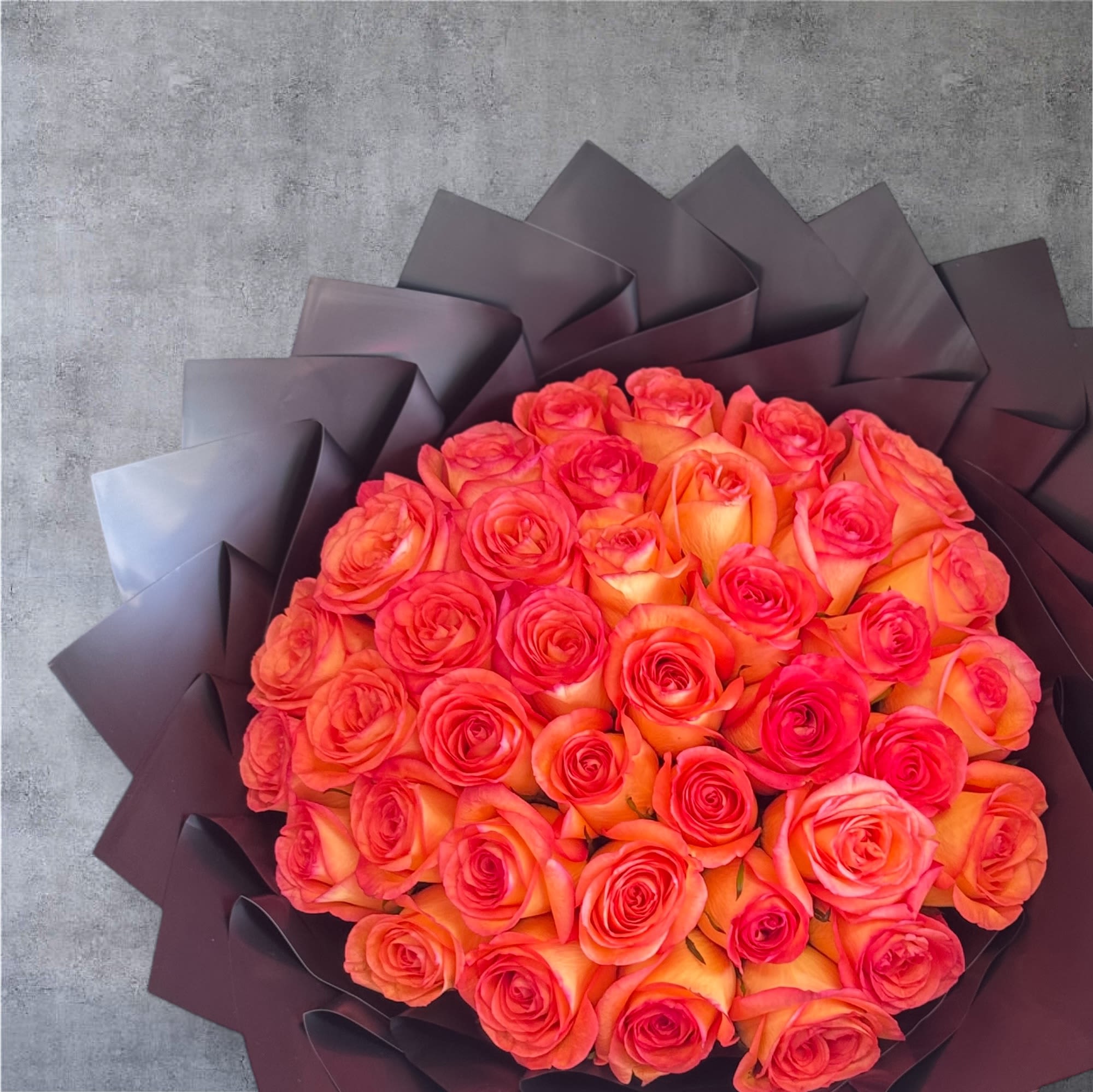 Round bouquet of bright orange and pink roses wrapped in dark folded paper.