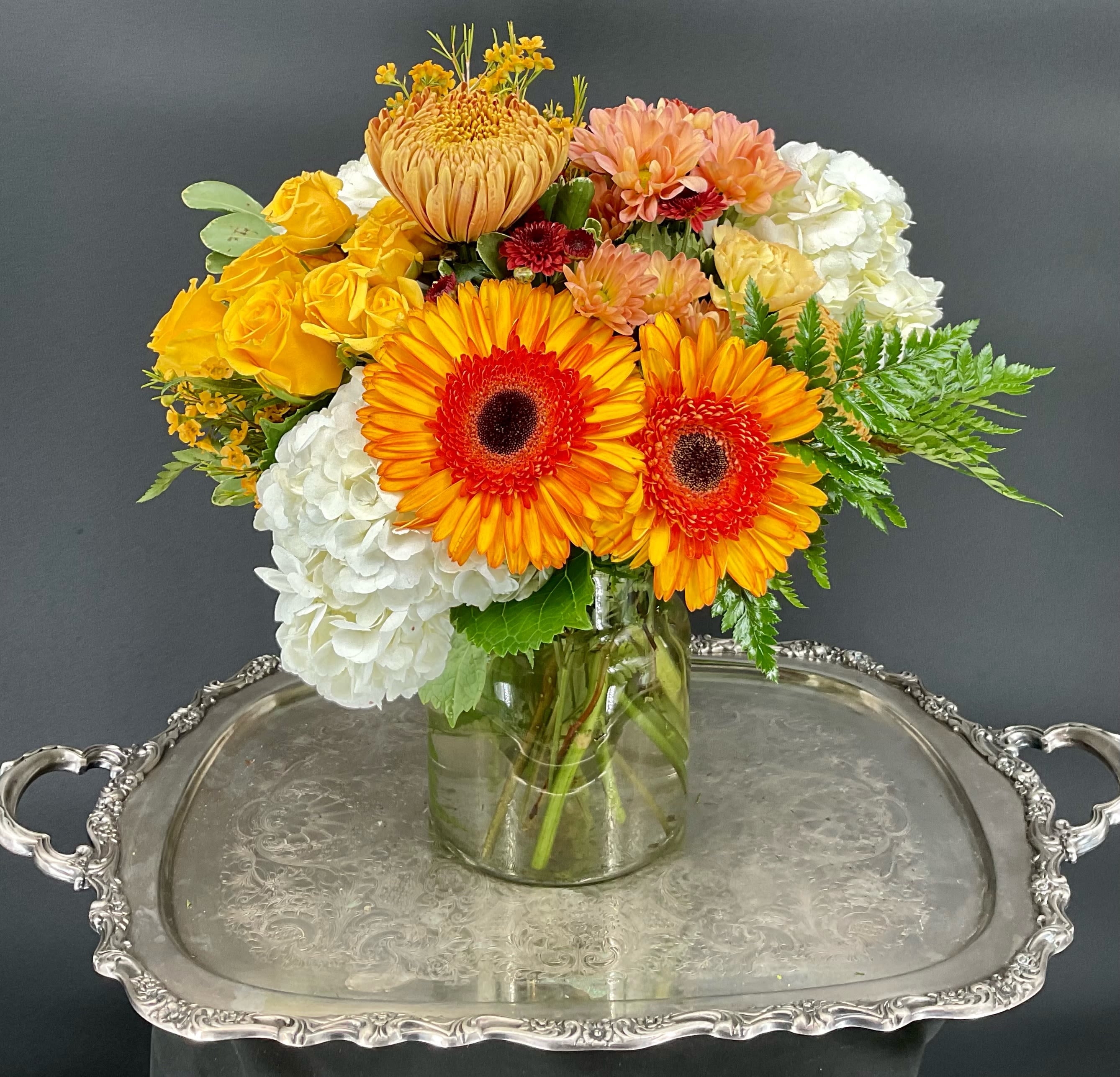 Bright mixed bouquet in a glass vase with orange gerberas, yellow roses, and white hydrangeas.