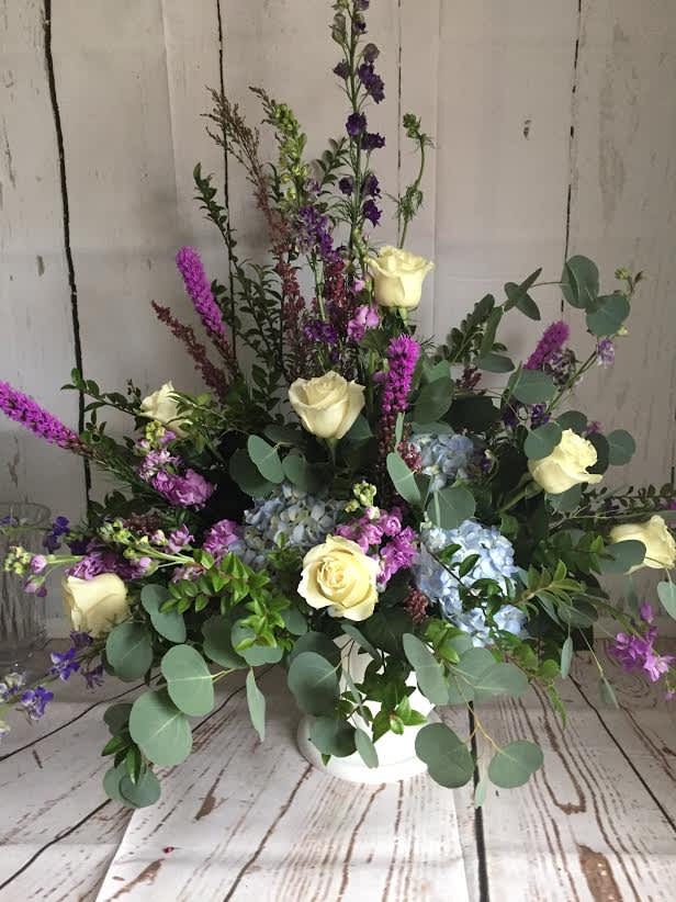 Arrangement of cream roses, blue hydrangeas, and purple spikes in a white vase