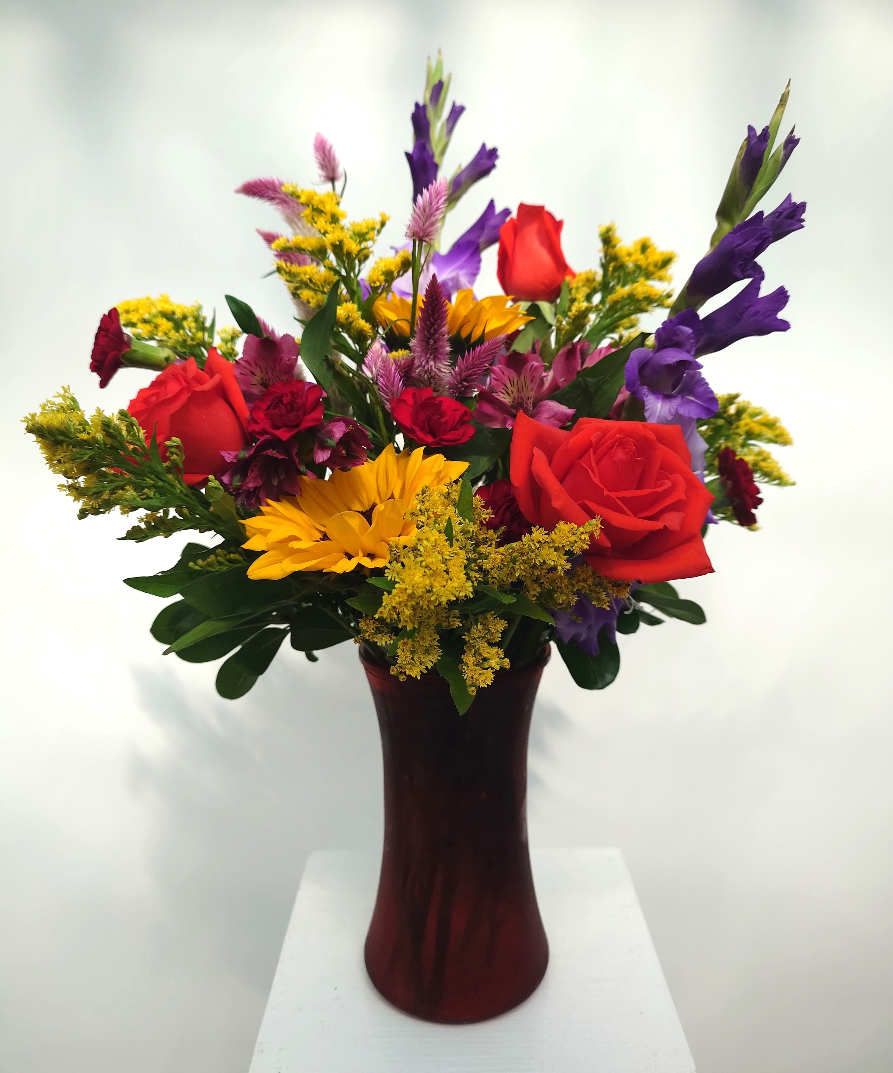 Mixed bouquet of red roses, yellow sunflowers, and purple flowers in a burgundy vase