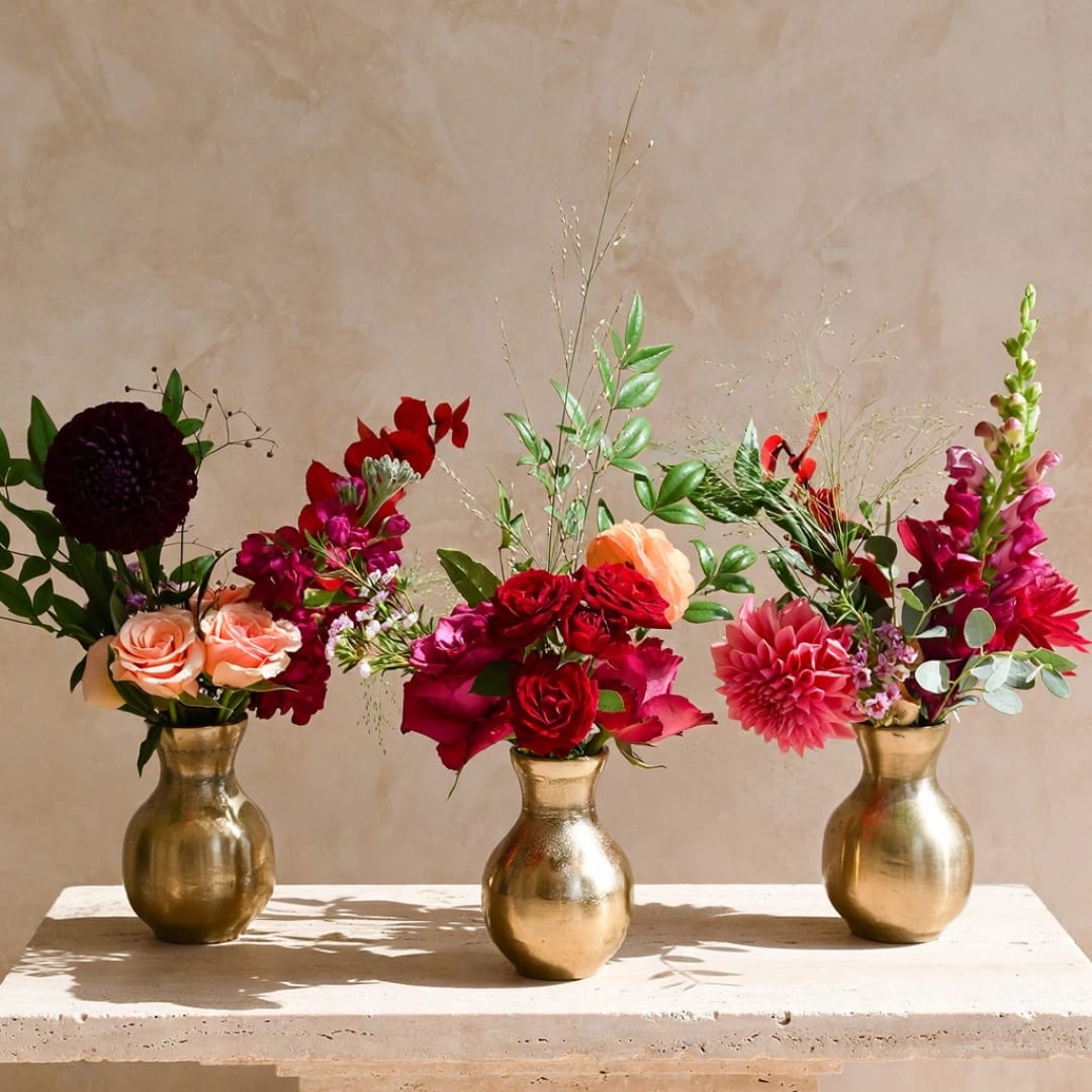 Three floral arrangements in gold vases with red, pink, and peach blooms