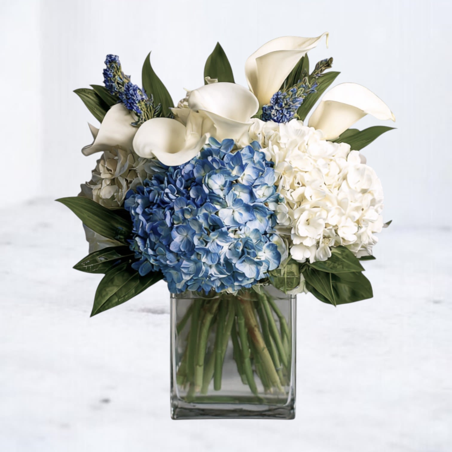 Low arrangement of blue and white hydrangeas with white calla lilies in a clear glass cube vase