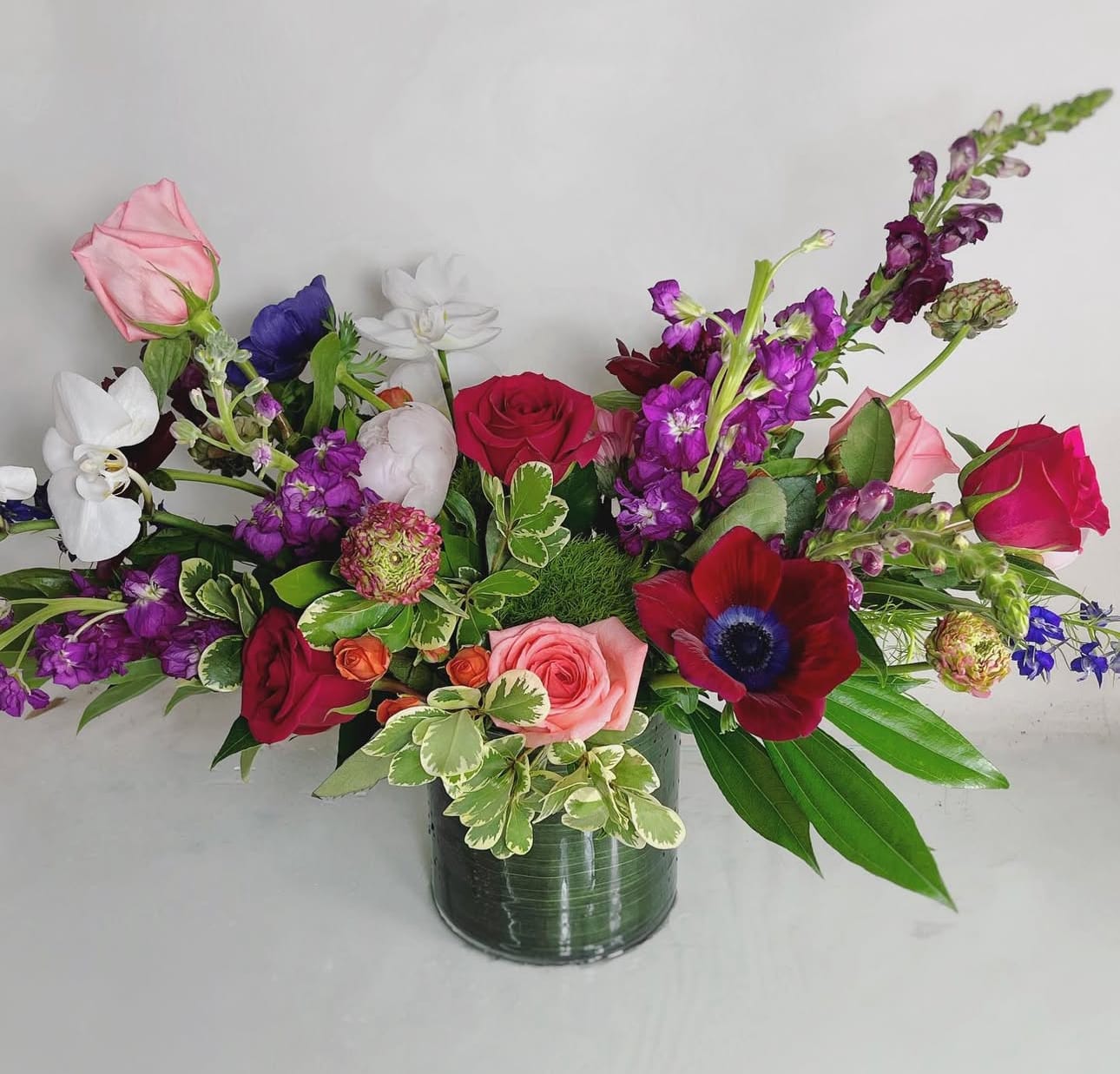 Petal Symphony by Twigs and Blooms - A breathtaking blend of passion and grace, this floral arrangement features a striking combination of red anemones, white orchids, stock, and roses.  The red anemones add a bold and vibrant touch, symbolizing love and anticipation, while white orchids bring an air of sophistication and purity. Stock flowers, with their delicate fragrance and lush texture, complement the bouquet with elegance, while classic roses—timeless symbols of beauty and devotion—tie the arrangement together in a harmonious display of color and emotion. Our Substitution Guidelines: Same or Higher Value: Any substituted flower will be of equal or greater value, ensuring your arrangement maintains its beauty and quality.  Similar Color Palette: We will replace flowers with alternatives that closely match the original color scheme and overall aesthetic of the arrangement.  Maintaining Design Integrity: The overall style, shape, and theme of the bouquet will be preserved, ensuring a look as close as possible to the original design.  Premium Flowers Always Used: If a specific bloom is unavailable, we will use the freshest premium flowers to create a stunning alternative.