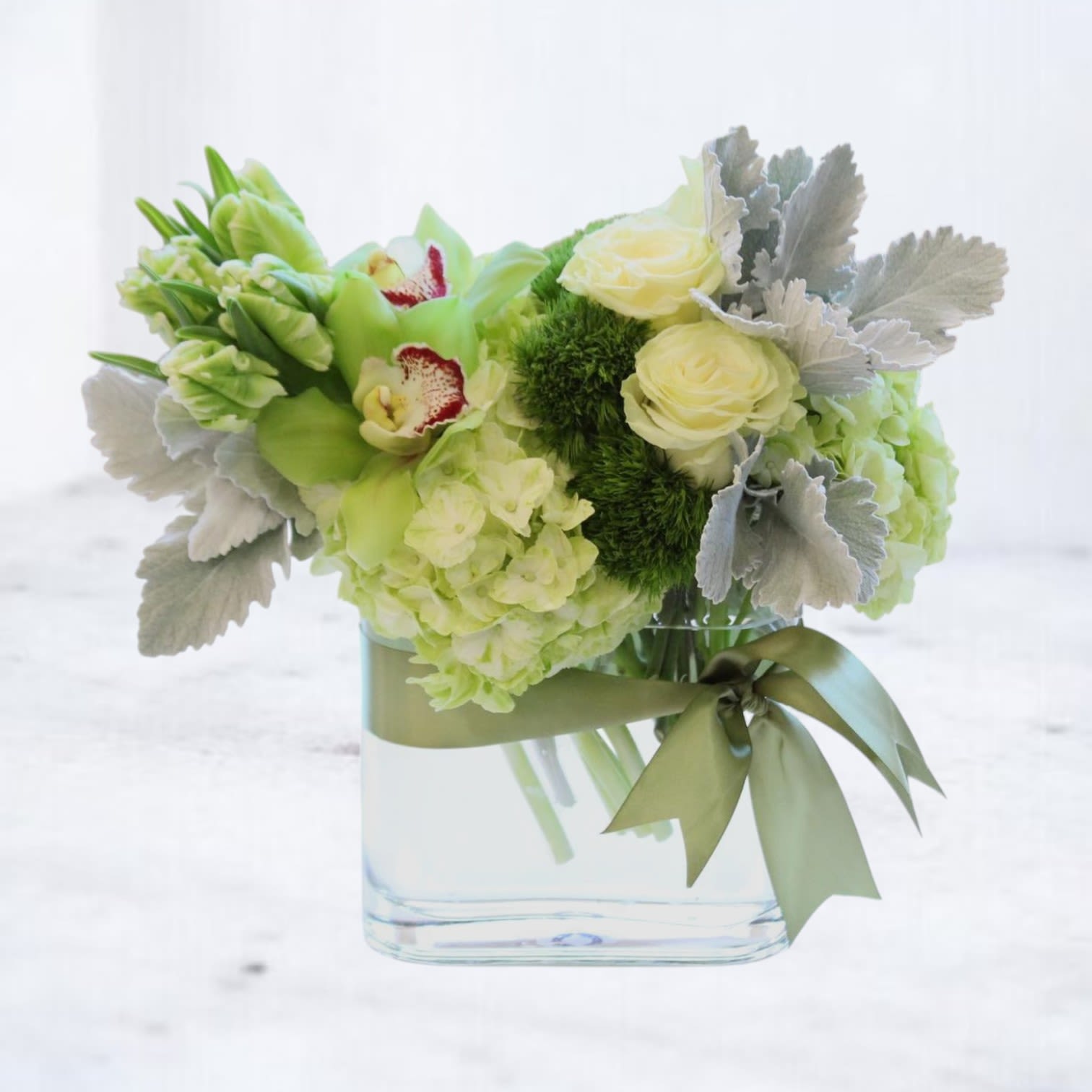 Silver Sage Garden - Silver Sage Garden is a refined and refreshing floral design that captures serene sophistication in soft green and silvery hues. This elegant arrangement features lush green hydrangea, pale ivory roses, cymbidium orchids, and mossy dianthus nestled among dusty miller and tulip buds. Tied with a satin sage ribbon and set in a sleek glass vessel, this bouquet is a modern nod to garden romance.  Perfect for corporate gifting, new beginnings, or sending a gentle gesture of love and support, this arrangement offers timeless beauty with contemporary flair.  Substitution Policy: Each arrangement is custom-designed with seasonal blooms. While we do our best to replicate the arrangement as shown, substitutions may be made based on seasonal availability. Any substitutions will maintain the integrity of the original design in color, style, and value.